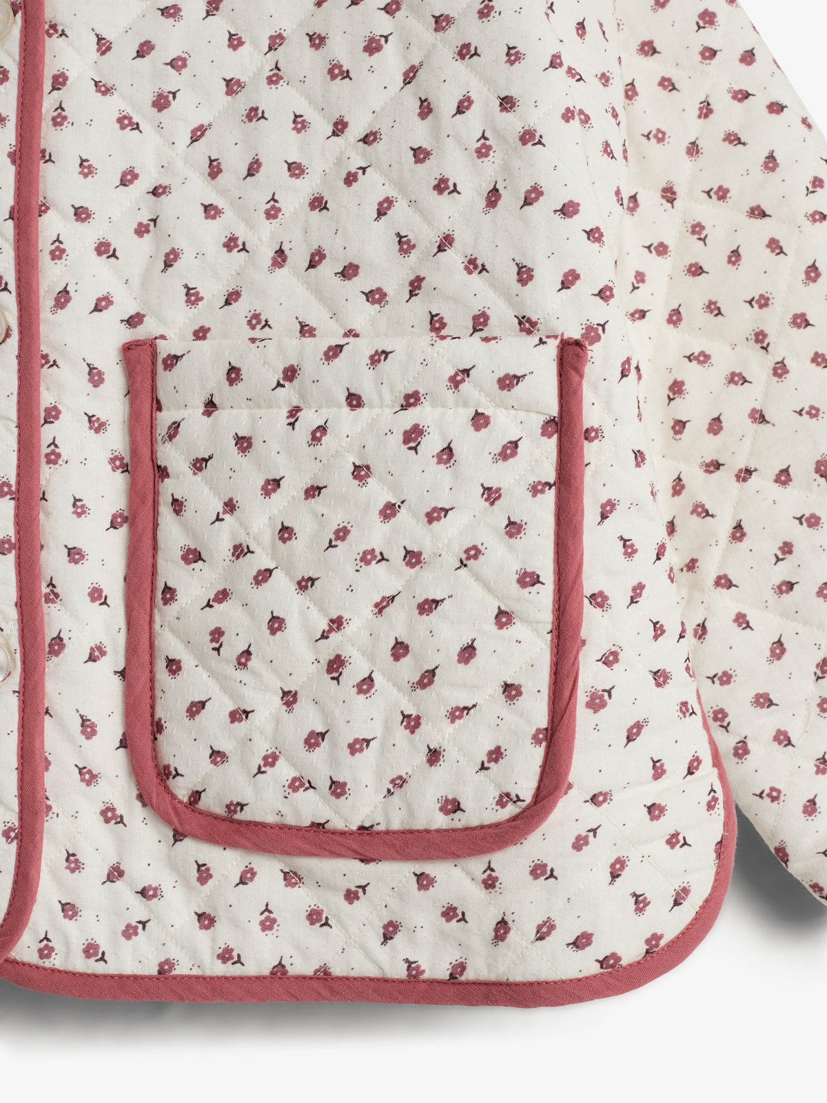 En hvid quiltet jakke med små, røde blomstermotiver over hele stoffet. Jakken har en firkantet lomme med matchende røde kanter, som fremhæver lommekanten i et rum.
