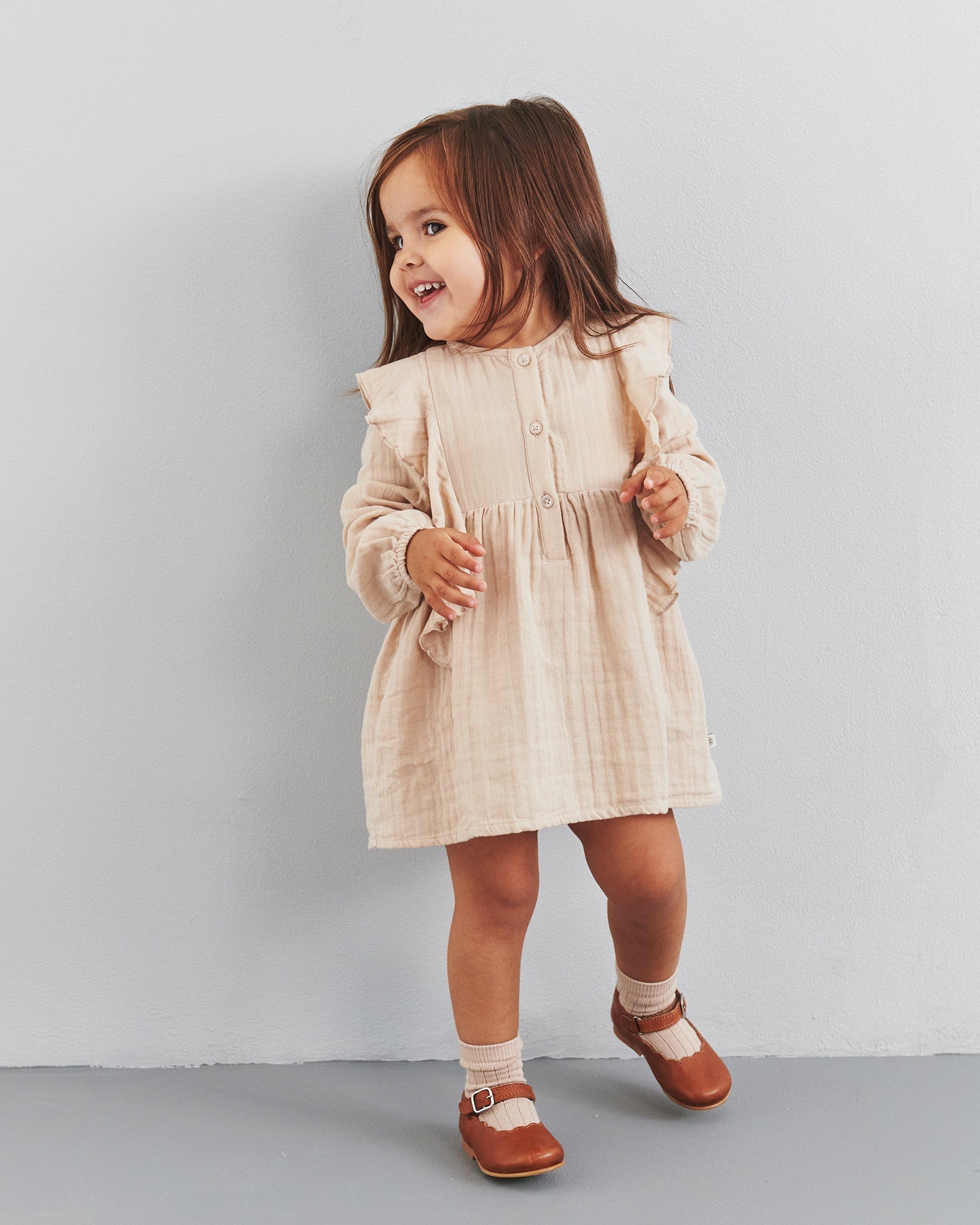 A small child wearing a beige dress and brown shoes stands and smiles against a plain, light gray wall.