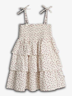 En hvid kjole med små røde blomster, flæselag og justerbare stropper med sløjfer, hænger enkeltvis mod en lys baggrund.