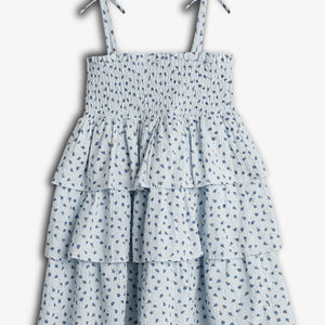 En blå, mønstret sommerkjole med bindebåndsstropper og flæser, hænger frit. Den har små blomsterdetaljer og en smockdetalje øverst, omgivet af en enkel, lys baggrund.