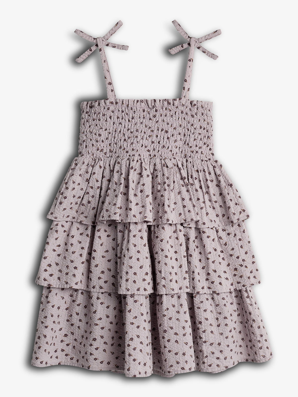 Kjole med spaghettistropper og bindebånd, dekoreret med små blomstermotiver, har flæselag. Baggrunden er neutral, hvilket fremhæver kjolens mønster og design.