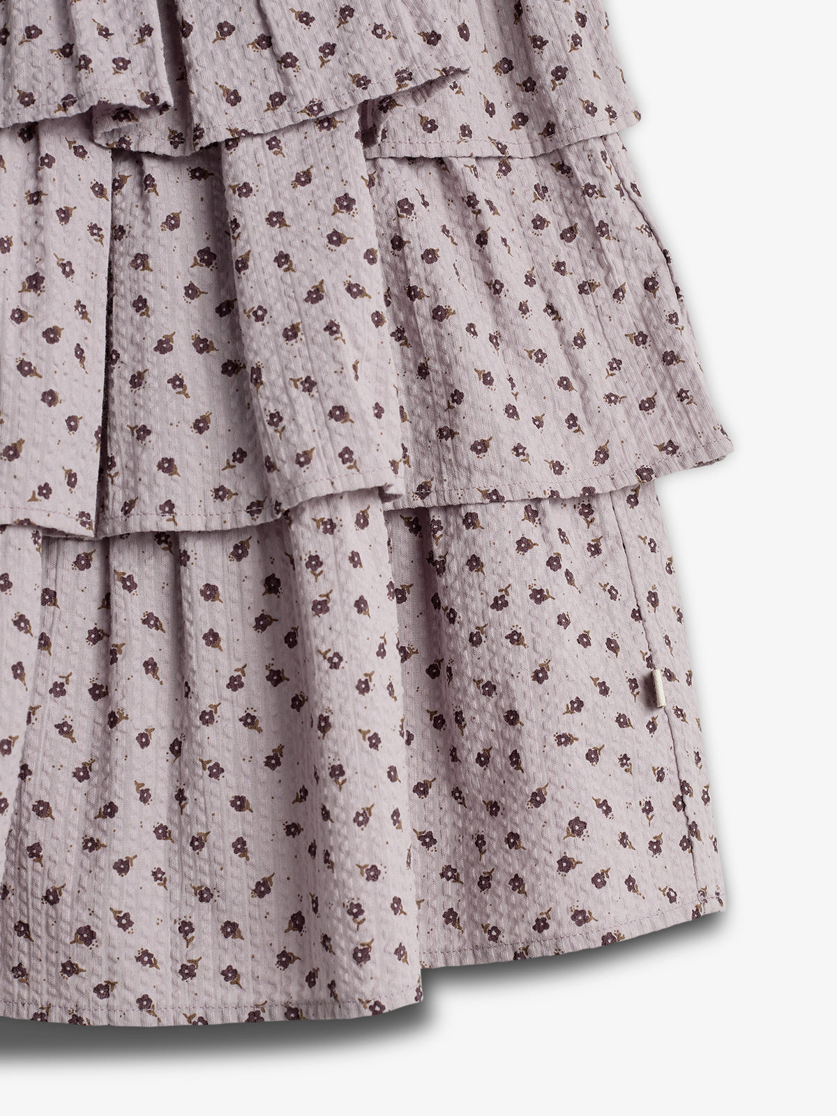 Kjole i flæselag med små blomstermønstre i mørkebrun på lys mauve baggrund. Stoffet er krøllet og lagdelt, hvilket giver en tekstureret og livlig effekt i neutral belysning.