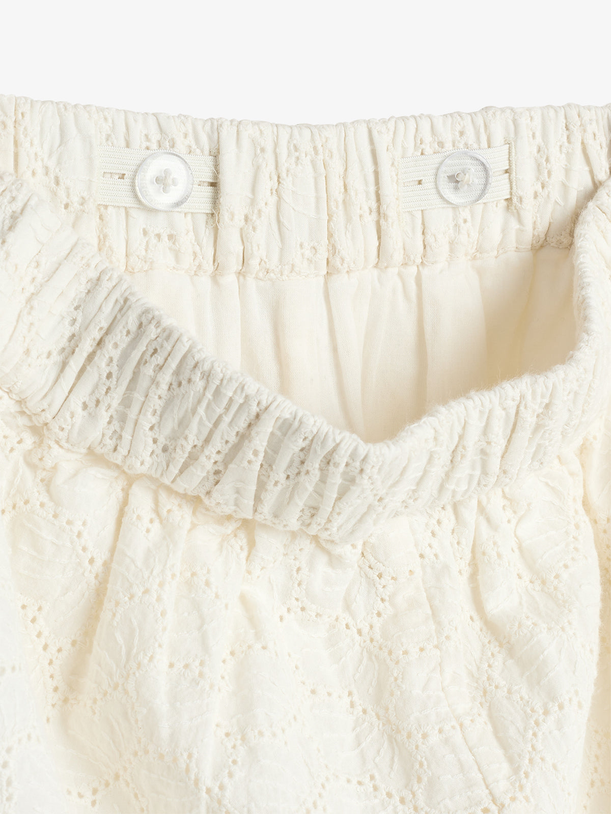Cream-colored lace skirt with a gathered, elastic waistband, featuring two buttons for adjustable fit. The fabric displays delicate floral patterns, creating a soft, elegant texture.