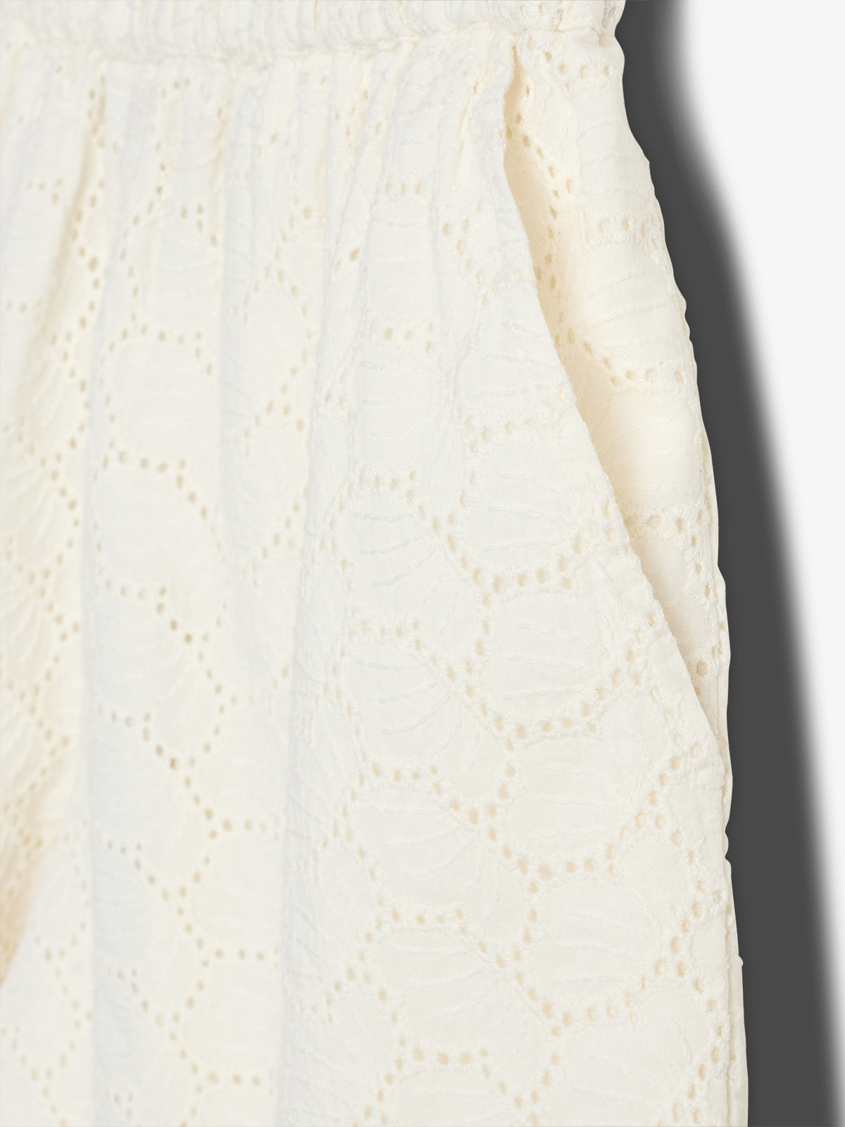 White fabric with a textured geometric pattern, including small holes, drapes softly. It features an elastic waistband, creating a gathered effect in a well-lit setting.