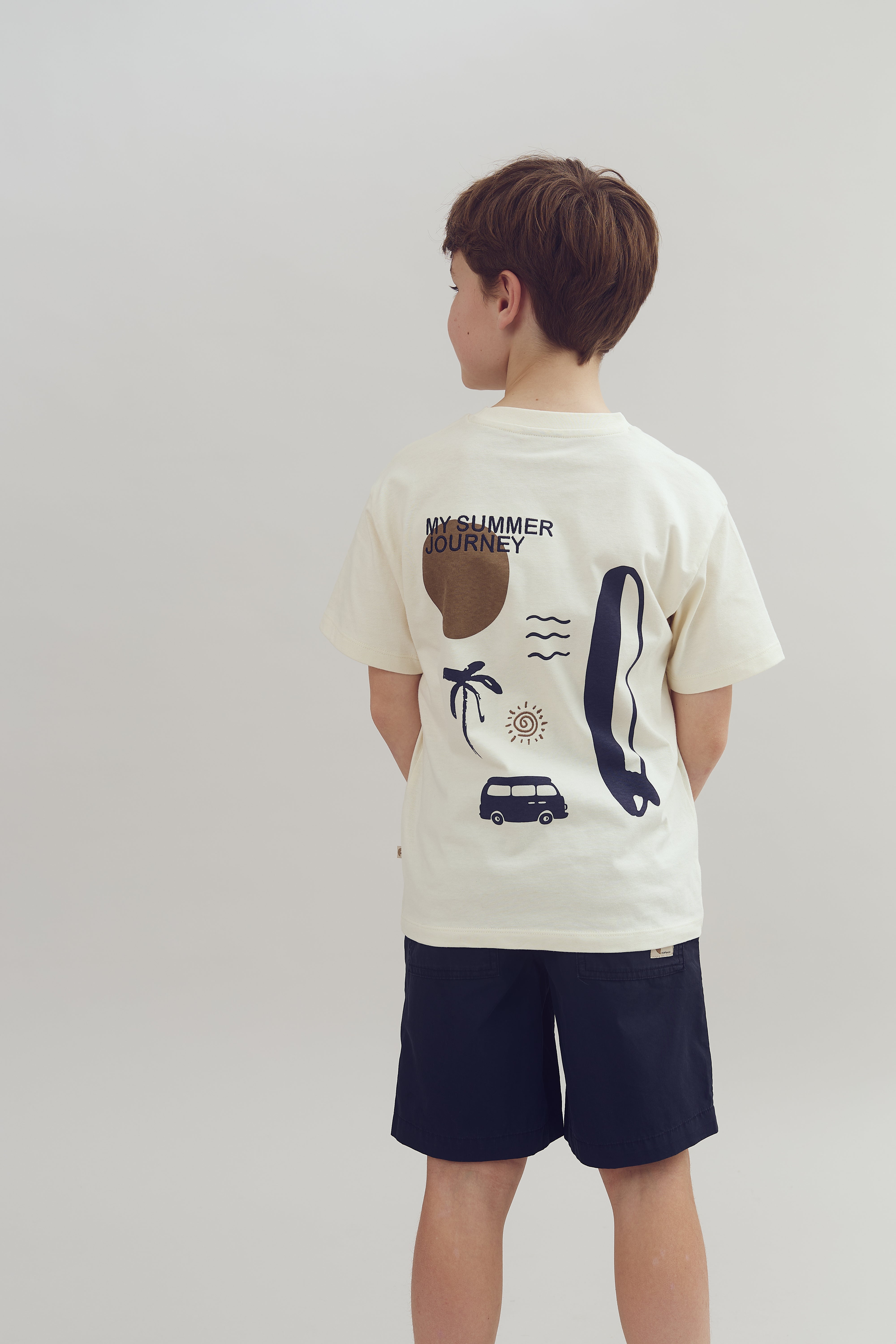 A child wearing a t-shirt with beach-themed illustrations, including a sun, palm tree, wave, van, and surfboard, stands in a neutral environment. Text reads "MY SUMMER JOURNEY."