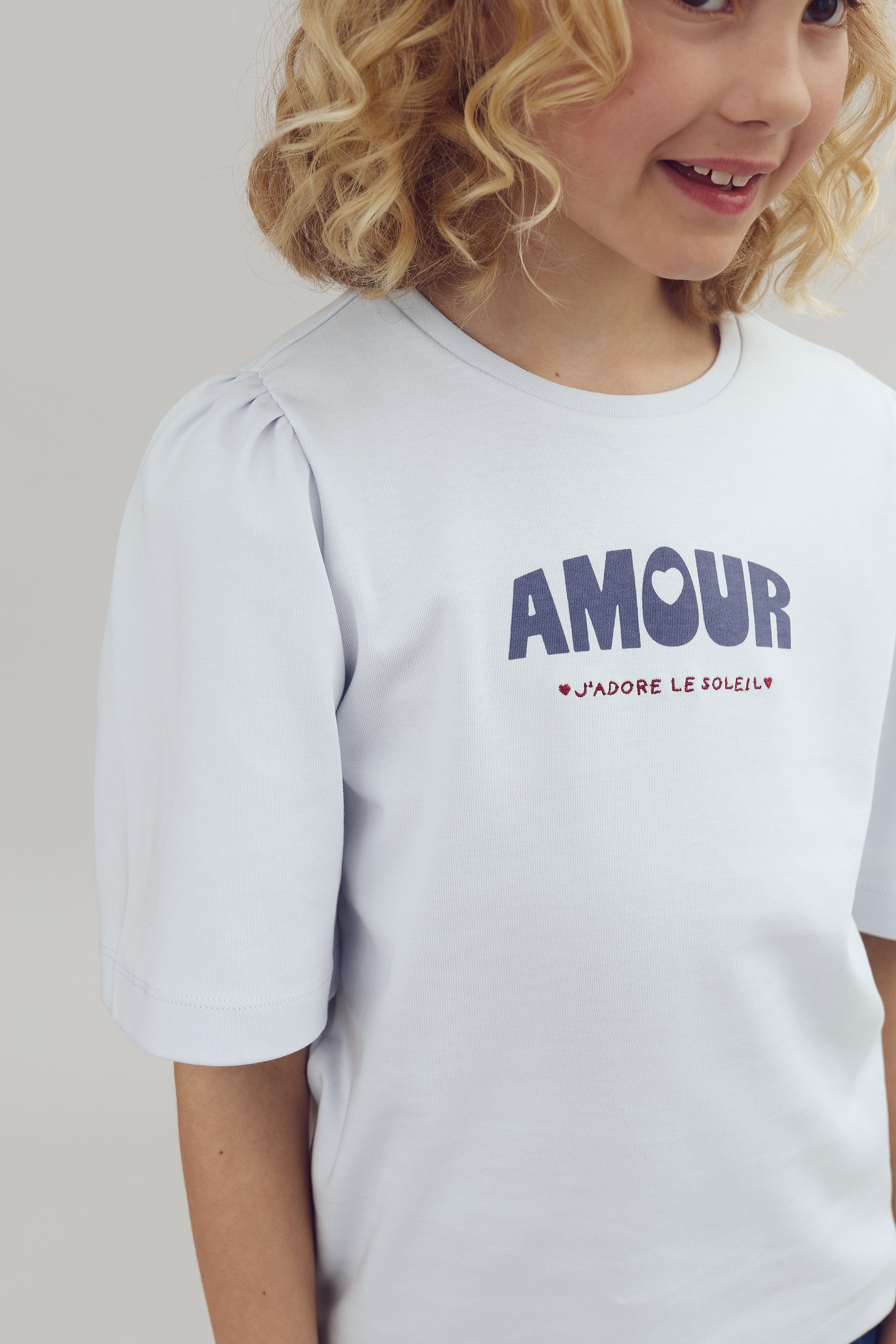 A child smiles while wearing a light blue shirt. The shirt features the text "AMOUR" and "J'ADORE LE SOLEIL" in bold letters. The background is plain.