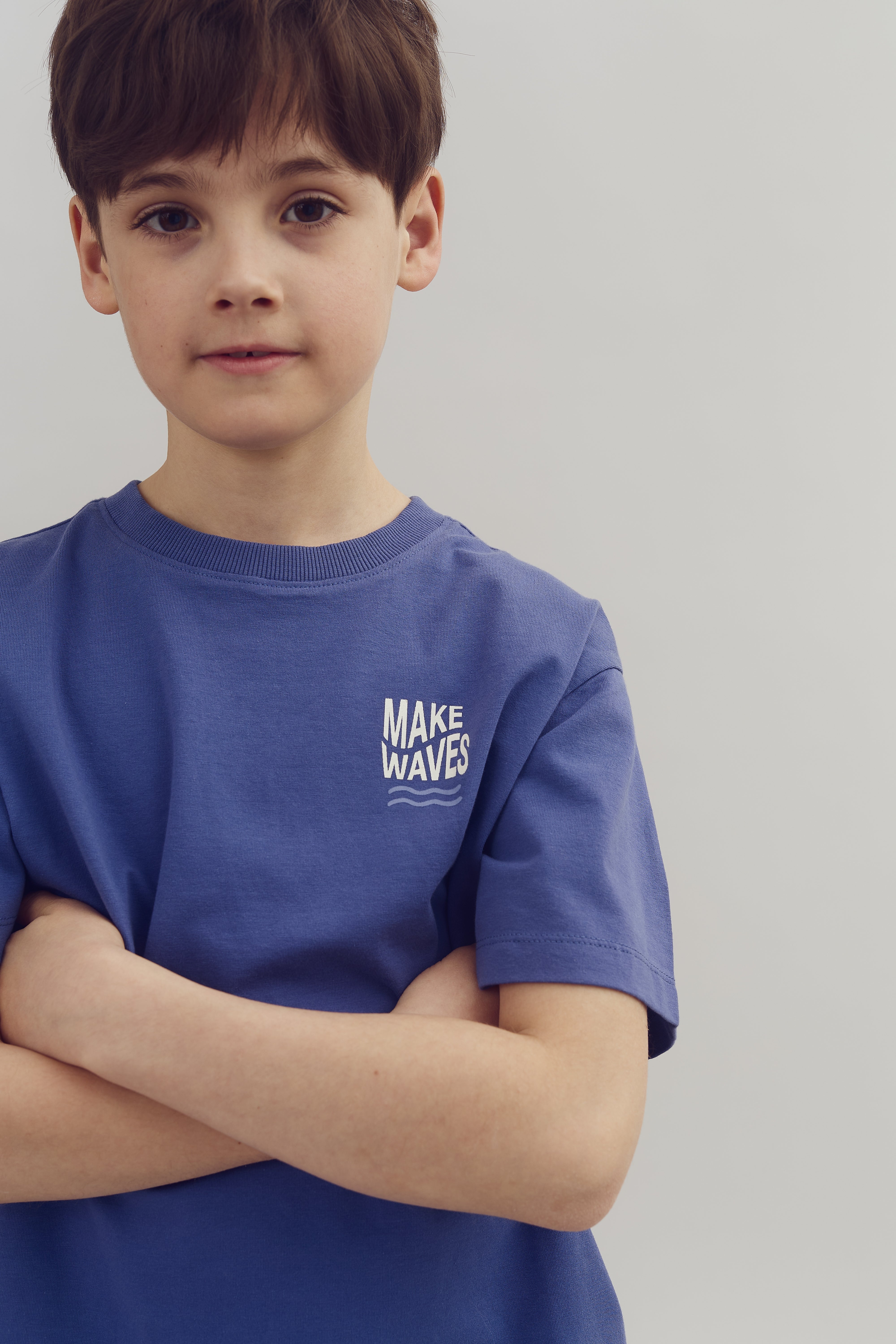 A child stands with arms crossed, wearing a blue shirt with "MAKE WAVES" text printed on it. The background is a plain, light-colored surface.