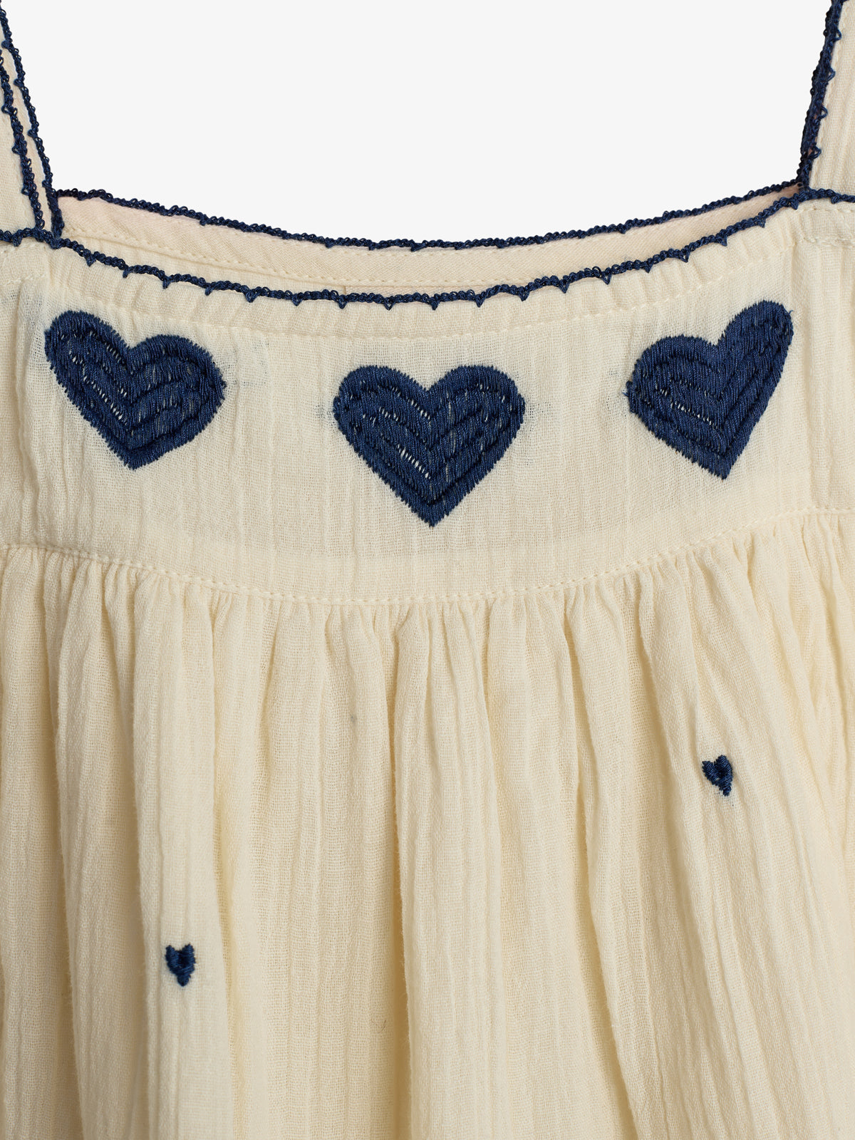 A cream-colored fabric with navy blue heart patterns embroidered along the top edge, featuring thin dark blue straps and gathered pleats below the embroidery, providing a textured appearance.