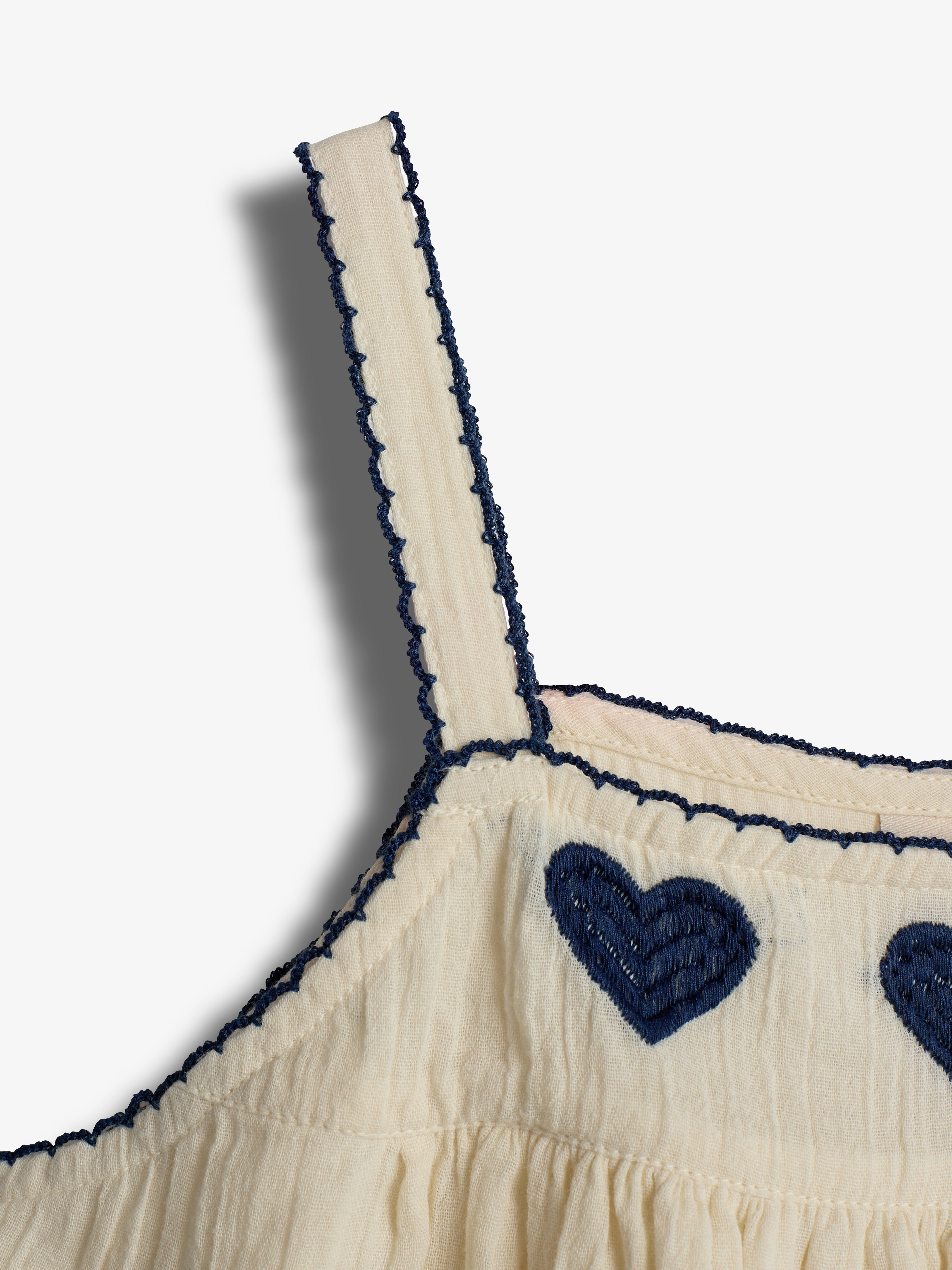 A cream-colored fabric strap with blue embroidered edges is attached to a garment featuring blue embroidered heart shapes; the background is a neutral, solid color.