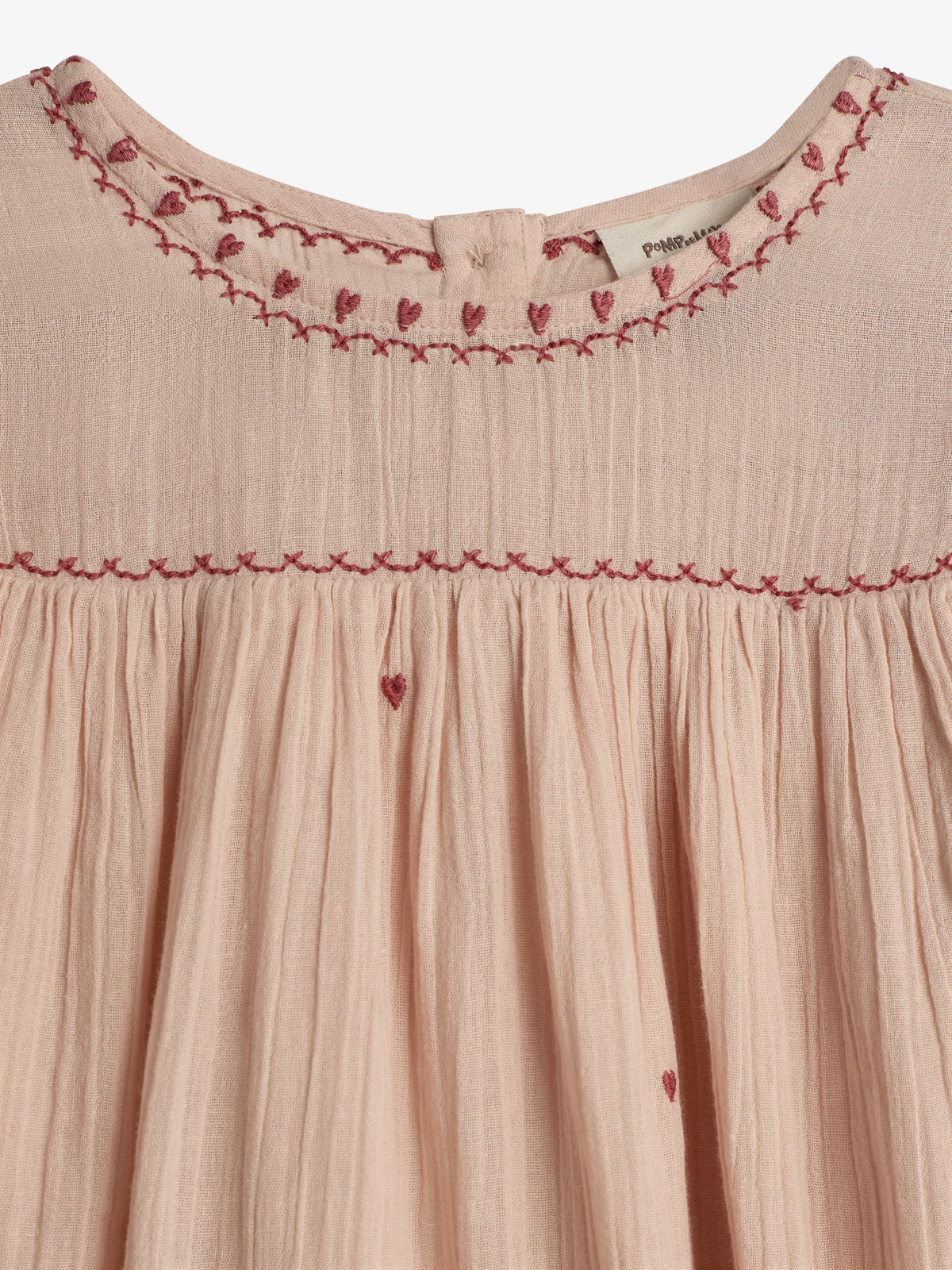 A pale pink dress features red heart embroidery along the neckline and pleated fabric. The label "Pompom" is visible near the top, adding subtle branding in a neutral setting.