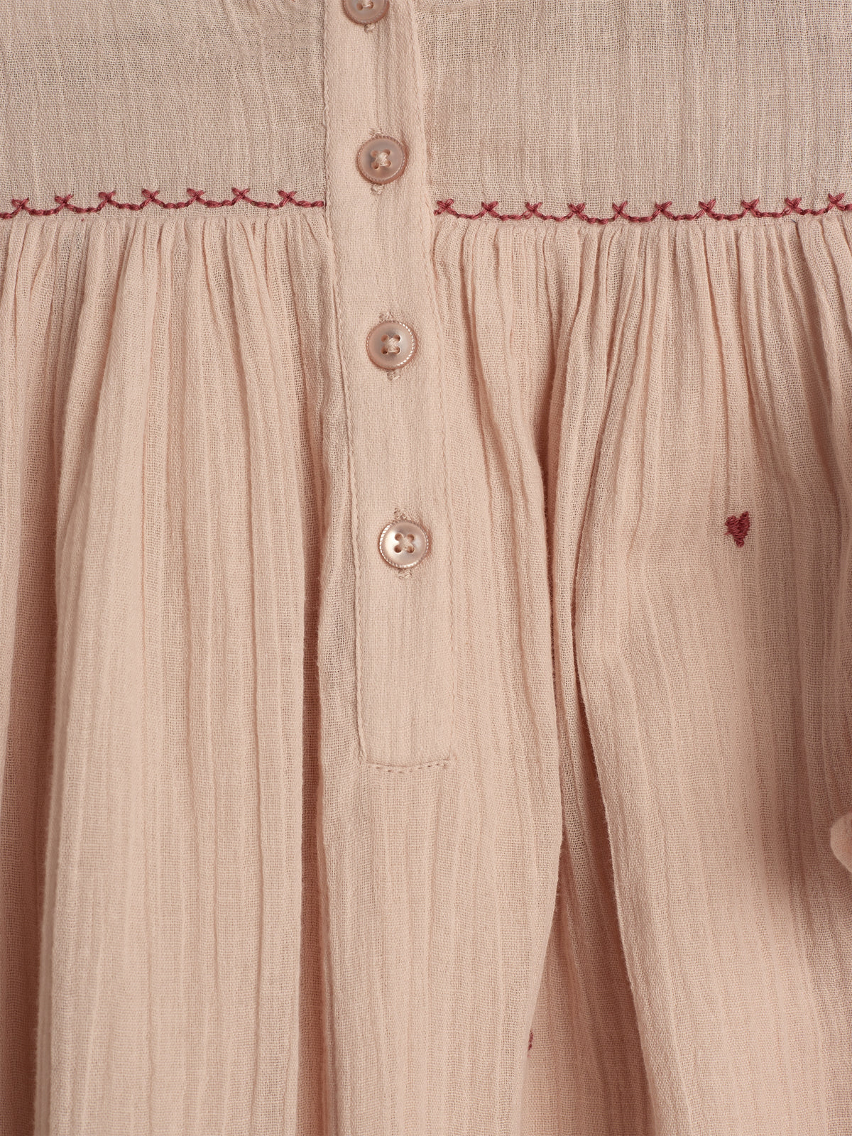 A beige fabric with a button-up design, adorned with red embroidered stitching and pleats, creates a textured appearance. Subtle red heart details accent the soft material.