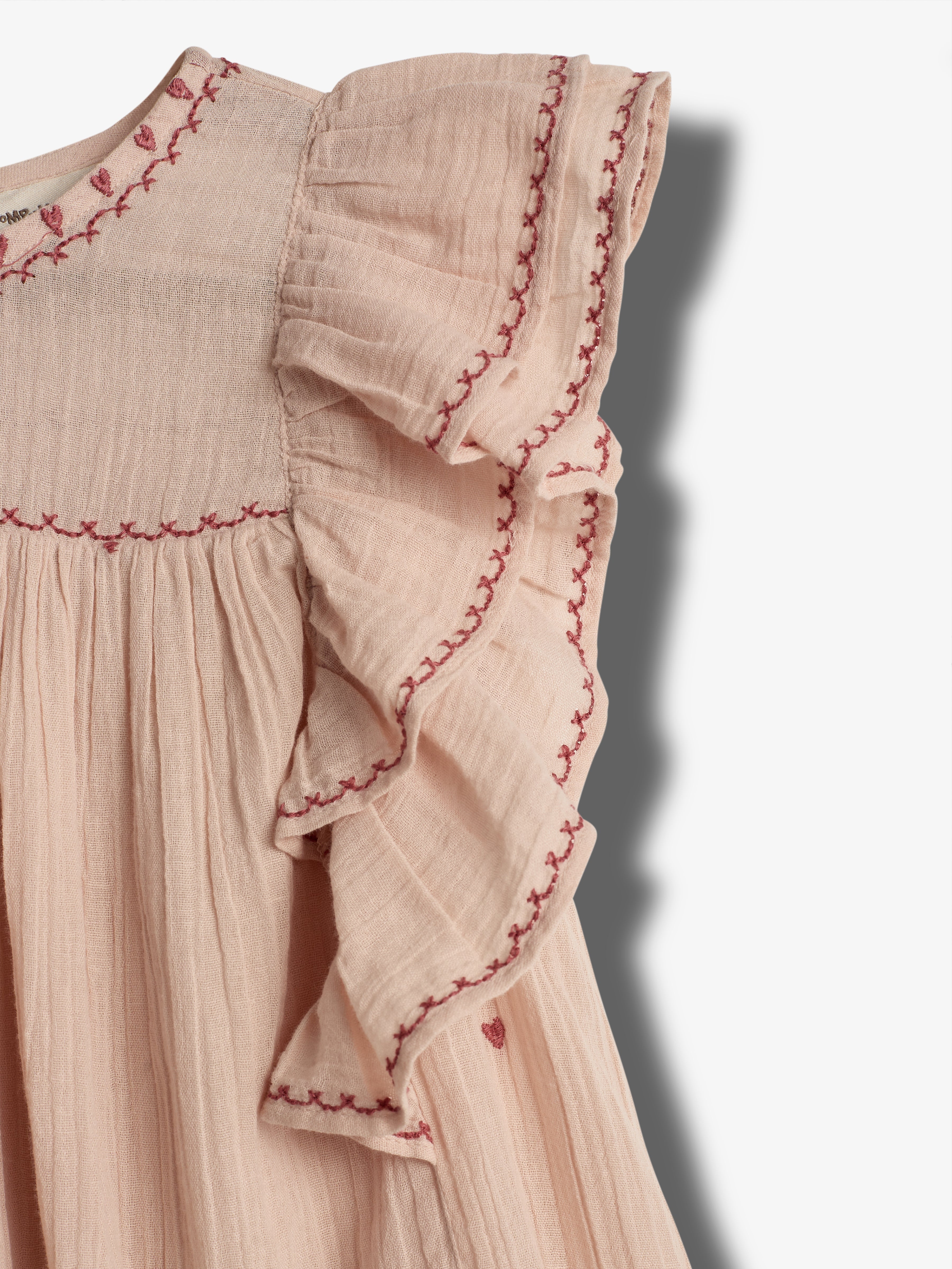 A pink, ruffled dress with embroidered red patterns decorates the neckline and sleeves. It hangs against a plain white background, showcasing its delicate, textured fabric.