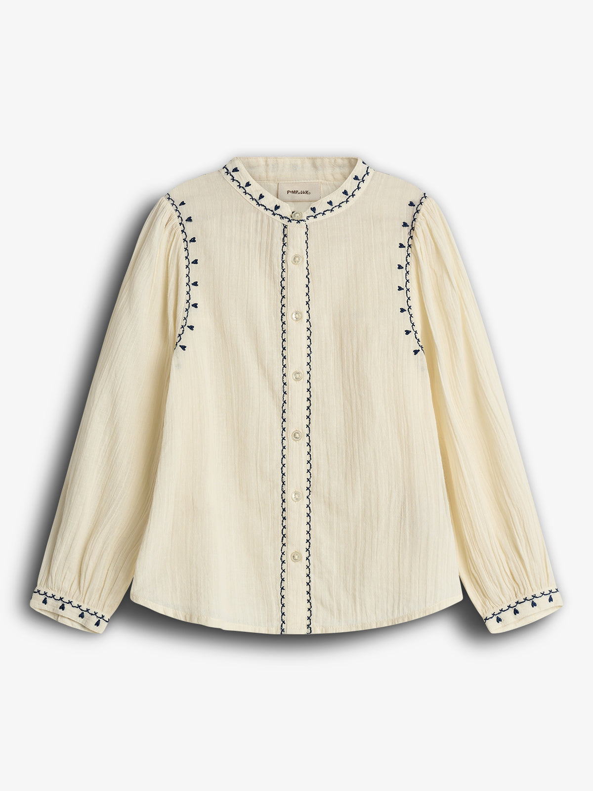 Cream-colored blouse with long sleeves, button closure, and navy blue embroidered details on the collar, cuffs, and along the button line. Set against a plain, white background.