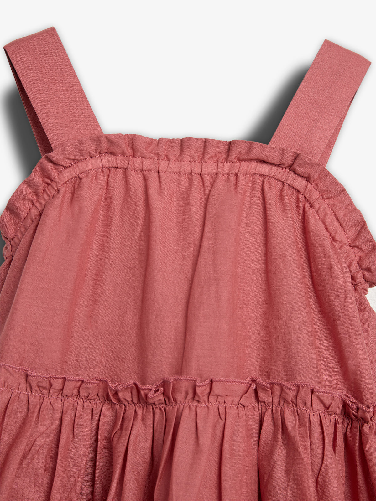 A pink sleeveless dress features ruffled detailing along the neckline and waist, with wide shoulder straps. The fabric appears soft and lightweight, set against a plain white background.