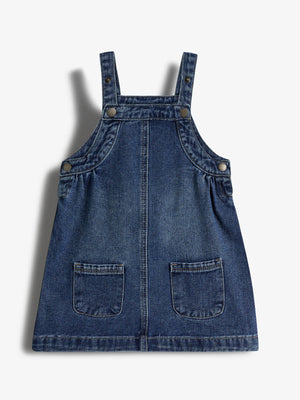 Blå denim selekjole med to forlommer, knappelukning ved stropperne og siderne. Kjolen er lagt fladt mod en hvid baggrund.