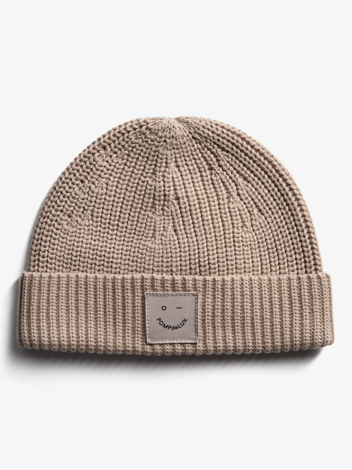 En beige strikhue med opsmøget kant har et mærke foran med et ansigt og teksten "POMPdeLUX." Ligger på en hvid baggrund.