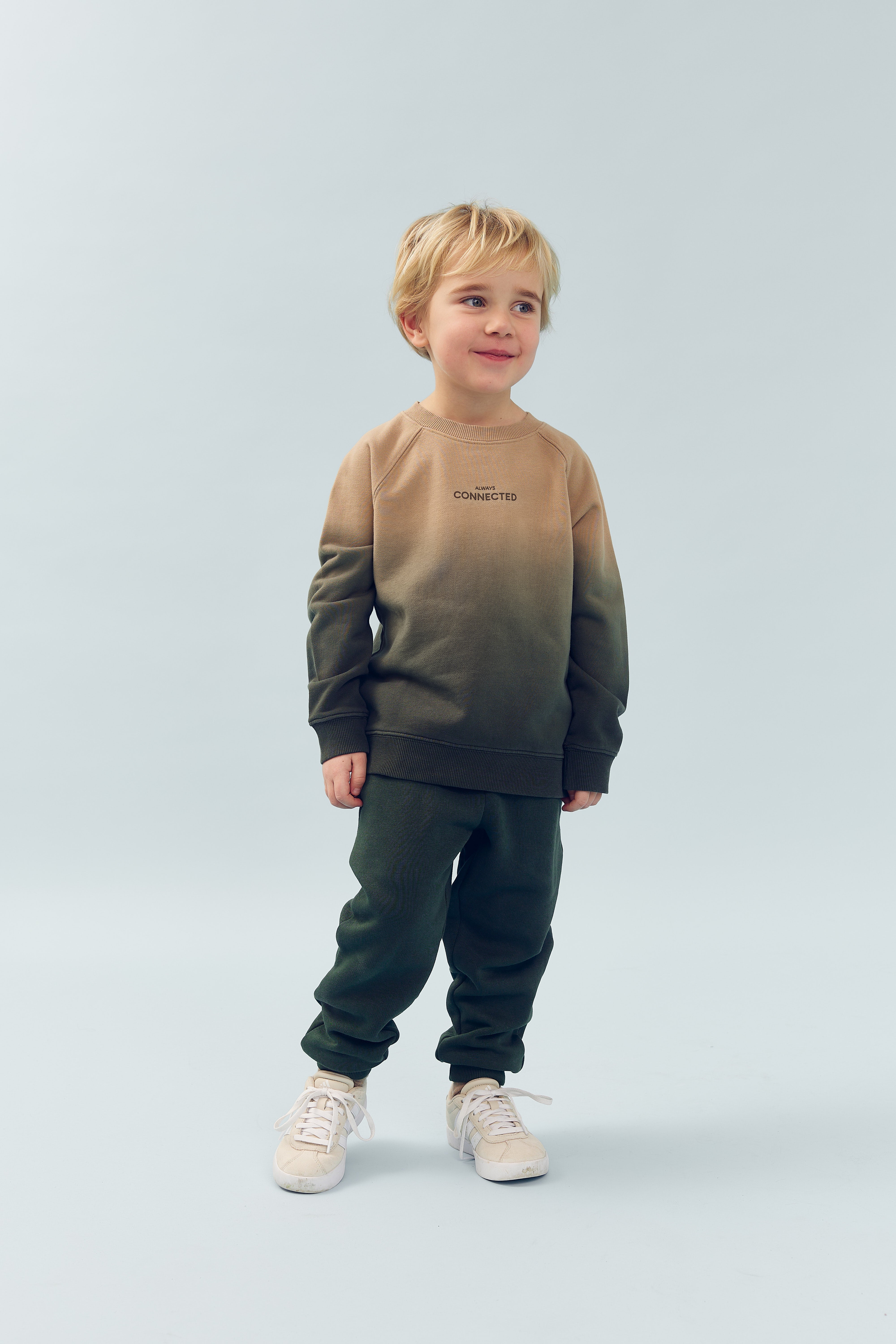 Child stands smiling in a light blue studio, wearing an ombre sweatshirt and pants with text "CONNECTED," and white sneakers.