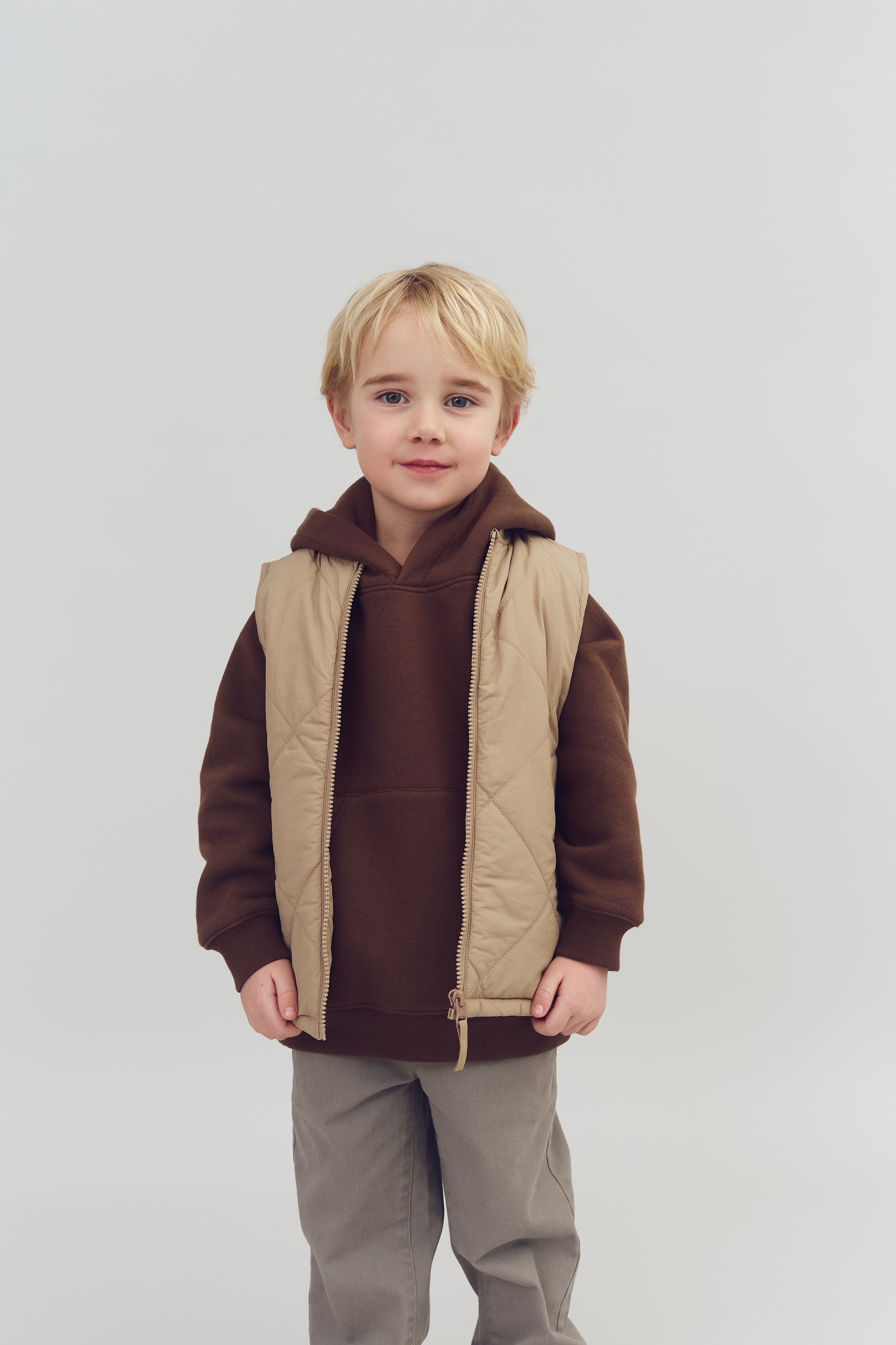 Barn står stille, iført brun hættetrøje og beige vest, med lysegråt bukser. Baggrunden er en ensfarvet, lys grå væg.