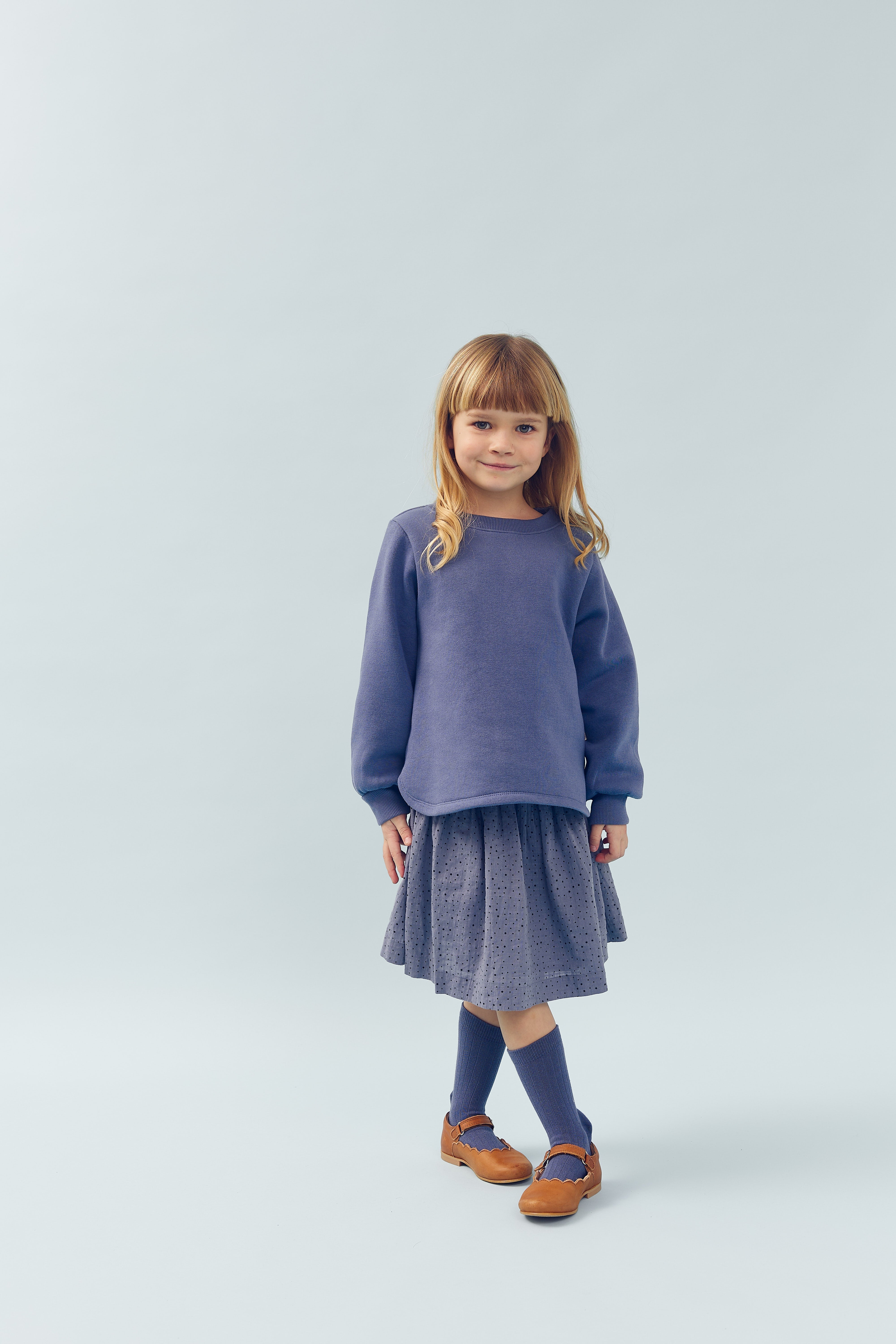 A child stands relaxed, wearing a purple sweater, polka-dot skirt, blue socks, and brown shoes against a plain light blue background.