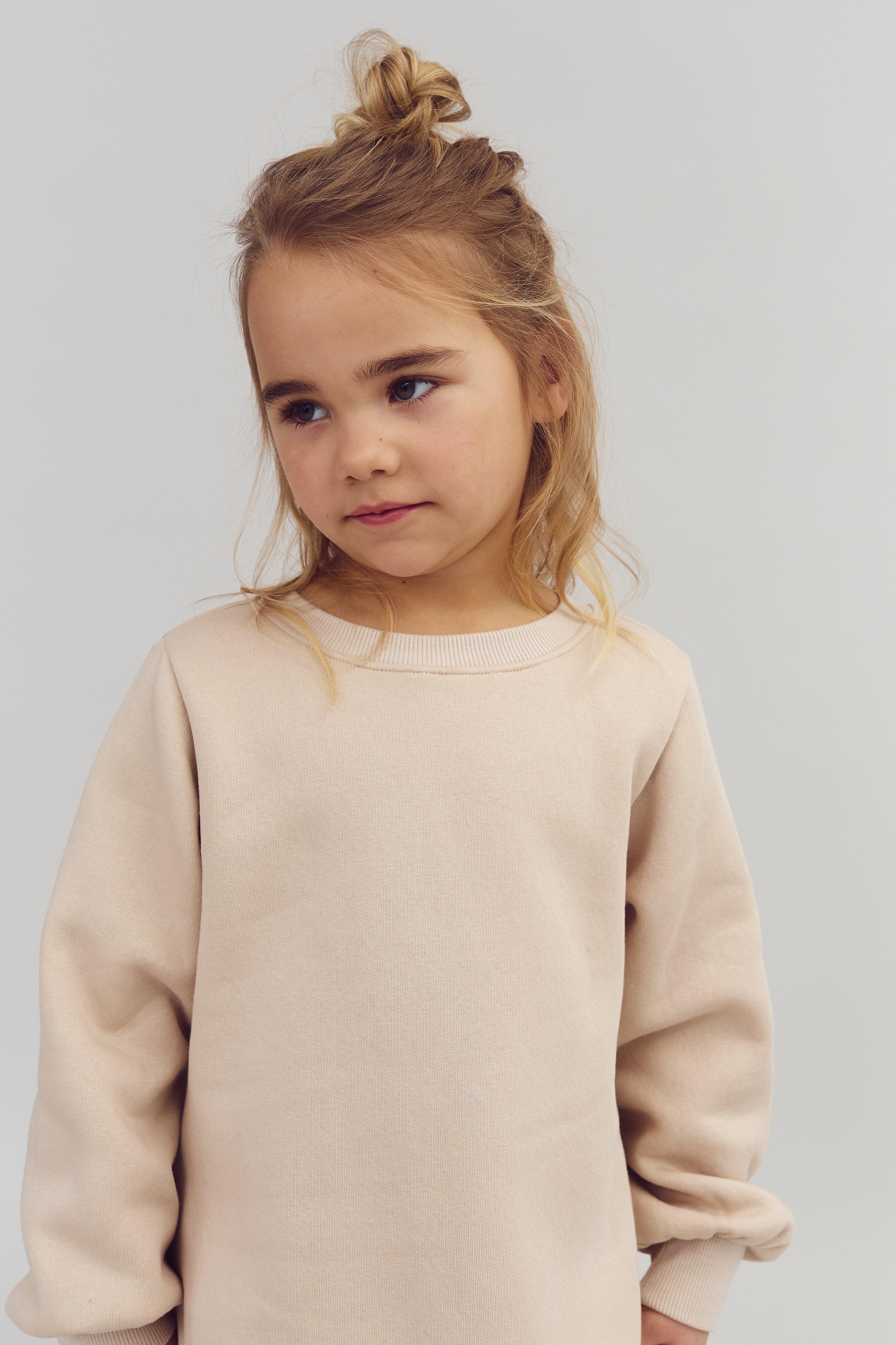 Pige i lyserød sweater ser opmærksom ud, står i et neutralt belyst rum. Hun har langt, lys hår i en knude, omgivet af en rolig atmosfære.
