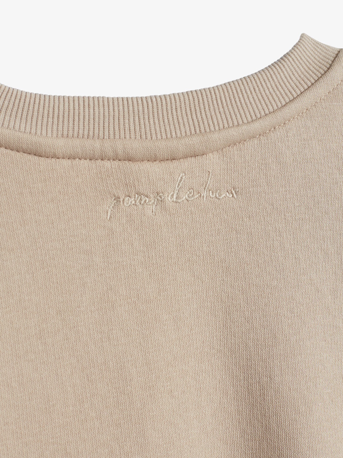 Beige stof med teksten "pomp de lux" broderet i hånden. Nærbillede af øverste del, med ribkant omkring halsudskæringen.
