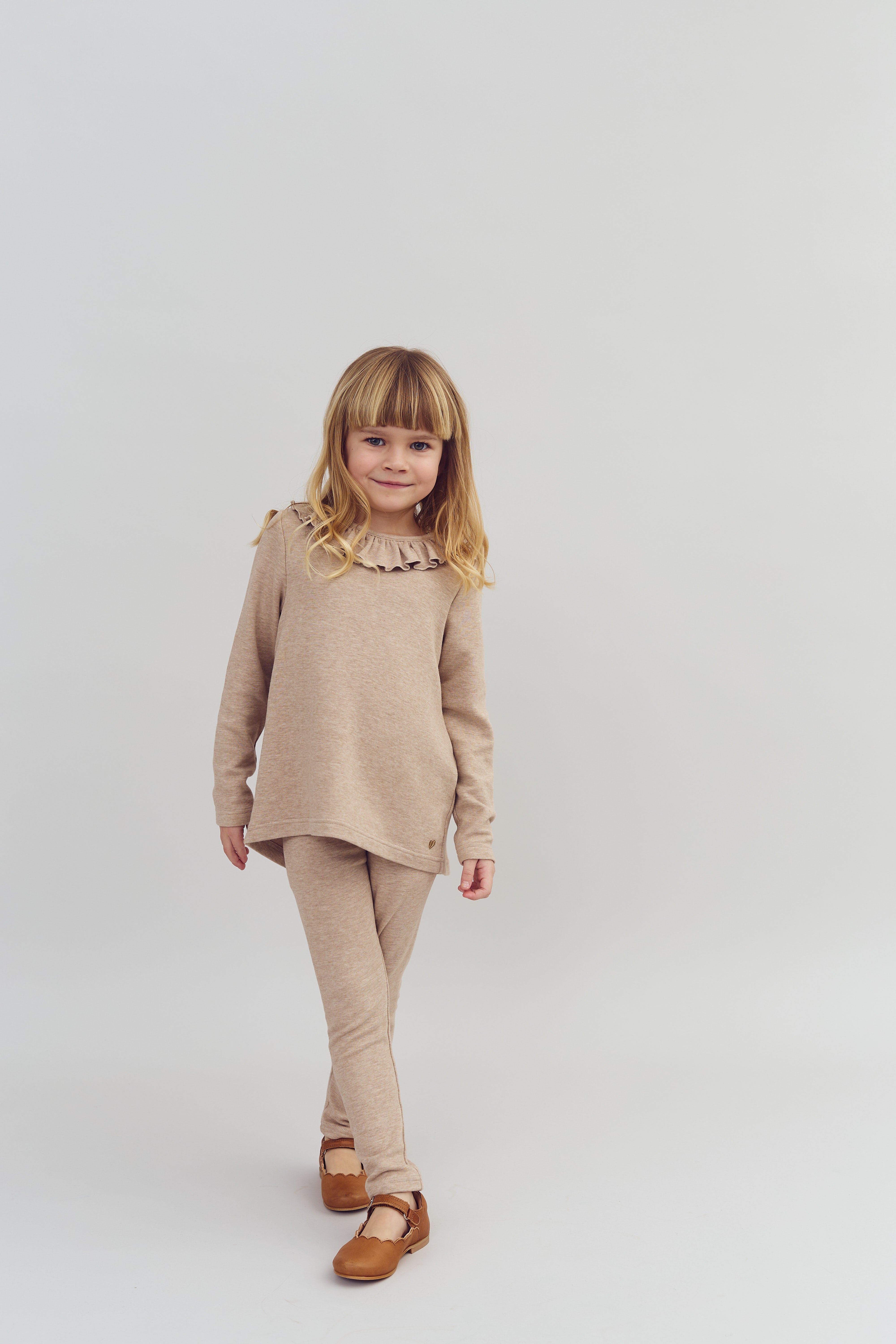 Et barn i en beige sweater og bukser smiler og går fremad i et neutralt rum med en lys baggrund. Barnet har lyst hår og brune sko.