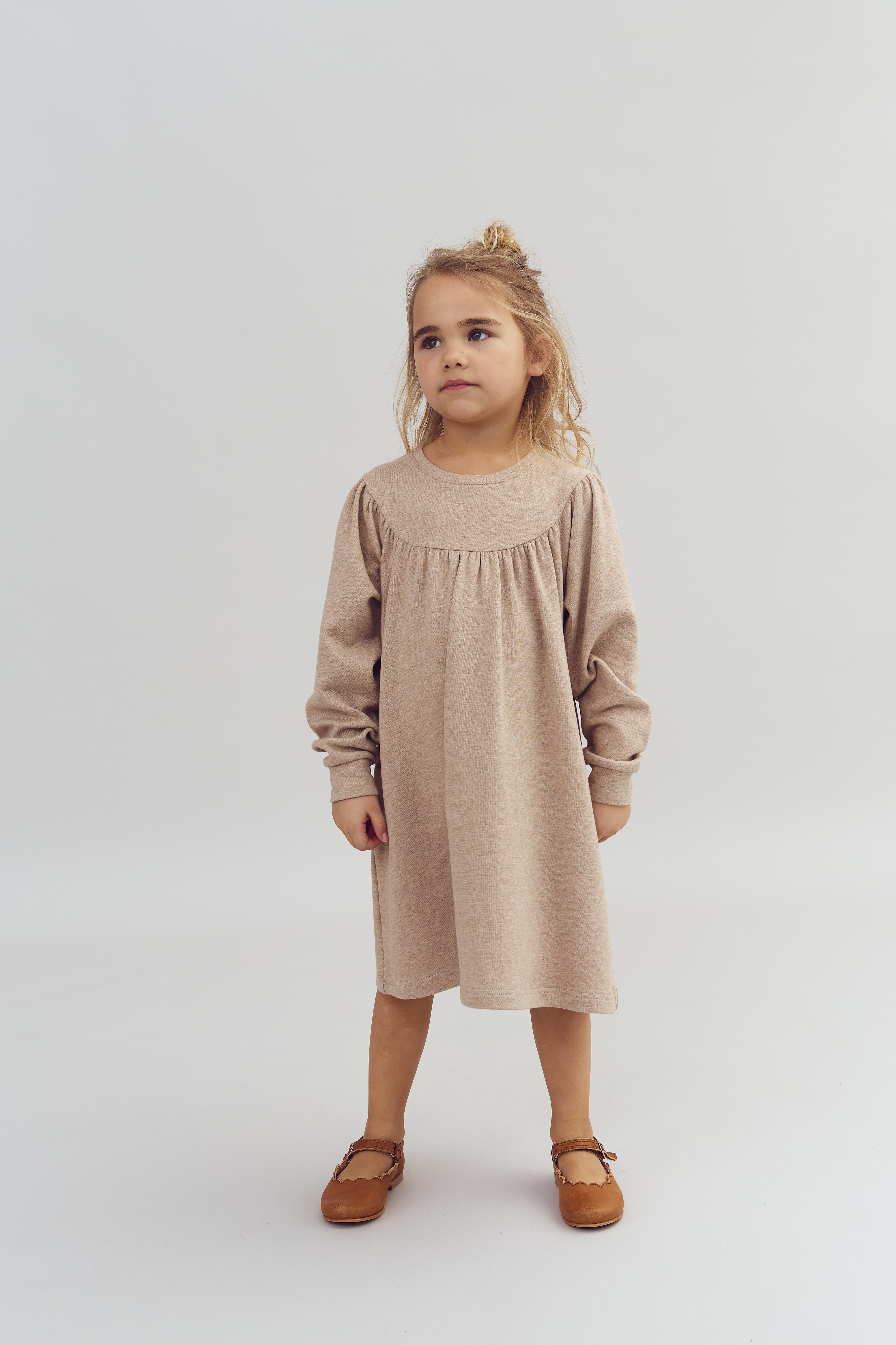 En lille pige står stille, iført en beige kjole og brune sko, i et lyst rum med en ensfarvet baggrund.