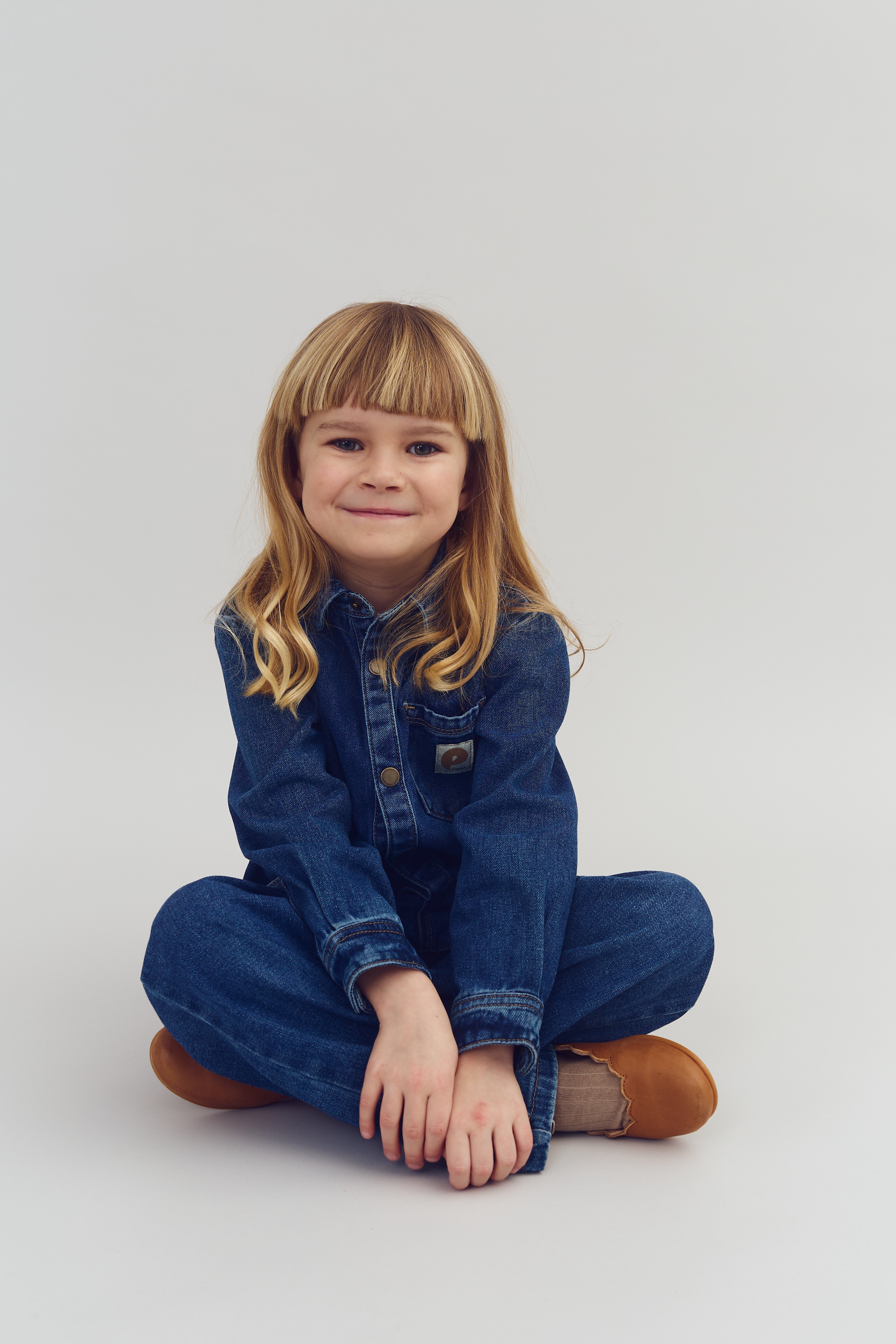 Et barn iført en denimdragt sidder krydsbenet på gulvet og smiler. Baggrunden er neutral og gråhvid, hvilket fremhæver barnets tøj og ansigt.