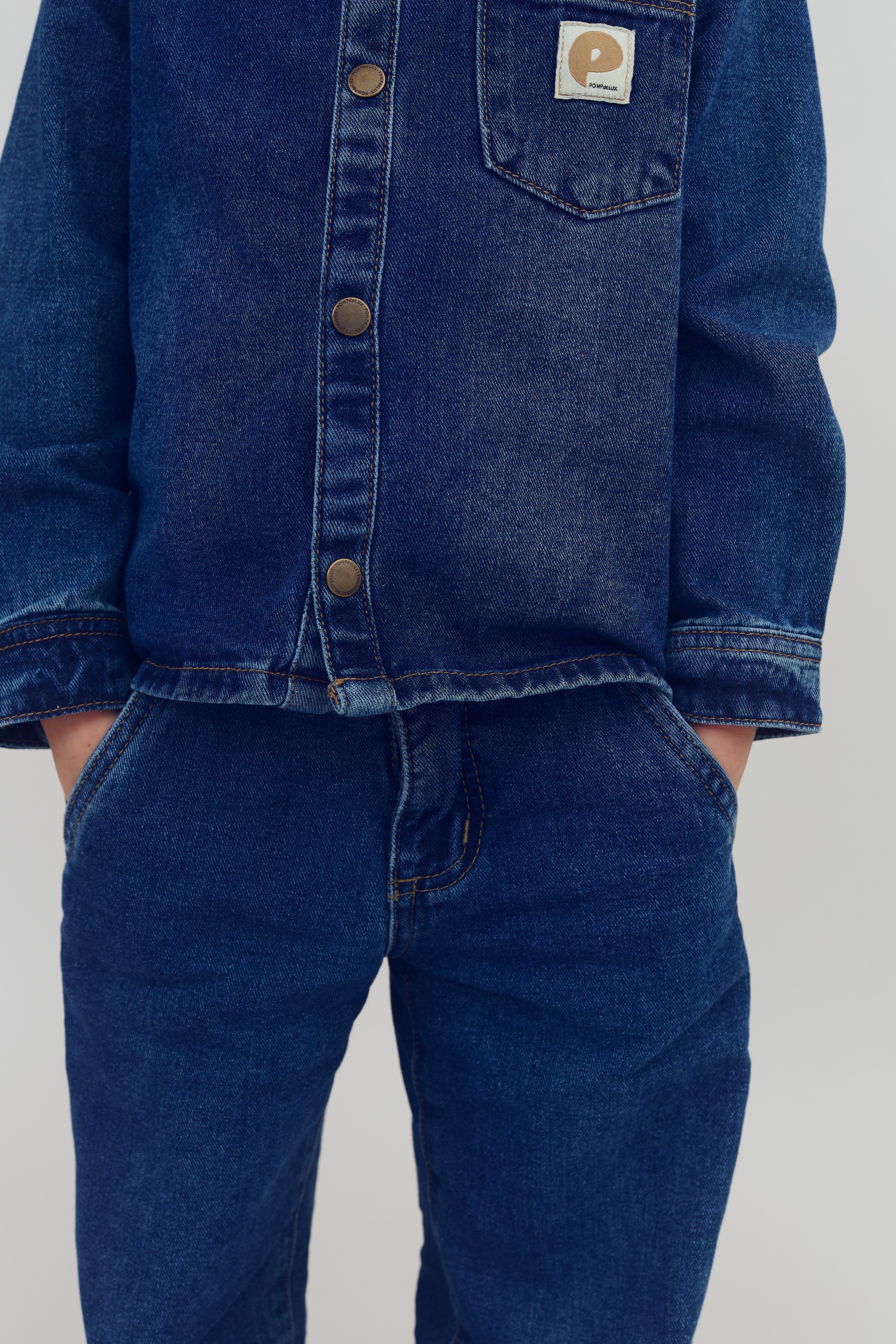 Jeansjakke med brune knapper bæres, hænderne i lommerne på matchende denimjeans. Patch med "P" og "Polarn O. Pyret" på brystlommen. Baggrund, lysegrå.