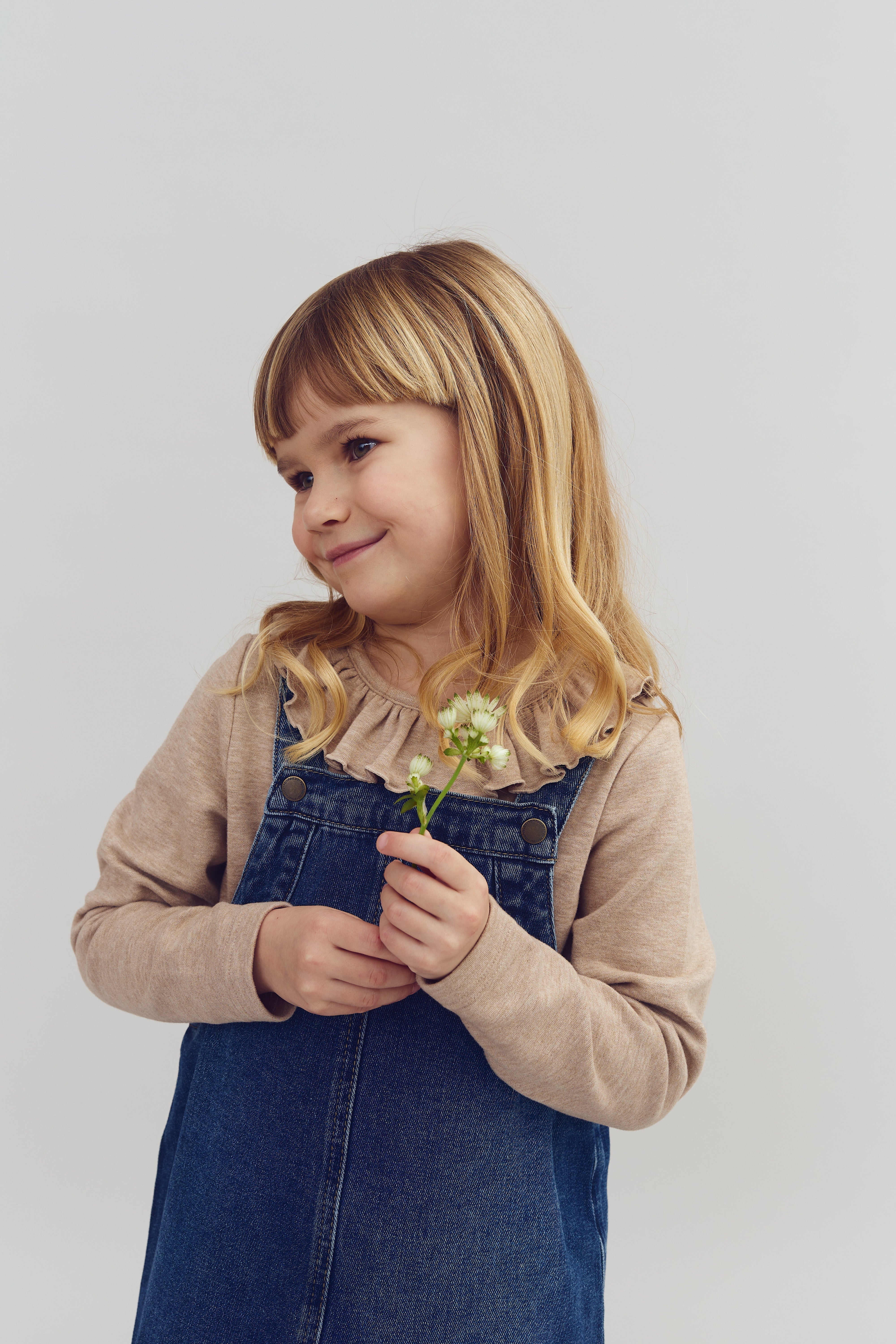 Barn holder en lille blomst og smiler; iført beige trøje og denimkjole, står mod neutral baggrund.