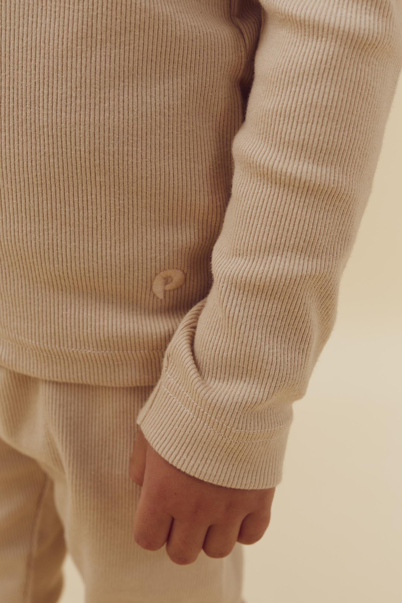 En hånd iført beige ribbet tøj, hvilende ved siden af kroppen, i en lys, ensfarvet baggrund.