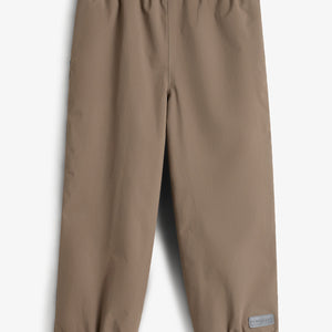 Bukser med elastisk talje og ankler, udført i en beige farve. Et lille rektangulært mærke nederst på det ene ben. Baggrunden er neutral hvid.