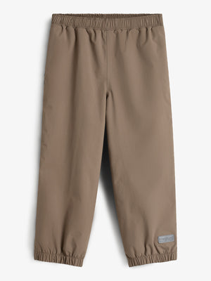 Bukser med elastisk talje og ankler, udført i en beige farve. Et lille rektangulært mærke nederst på det ene ben. Baggrunden er neutral hvid.