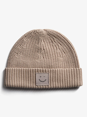 En strikket beige hue ligger fladt på en hvid baggrund. Der er et firkantet mærke foran med en simpel ansigtstegning og teksten "POMPdeLUX" under.