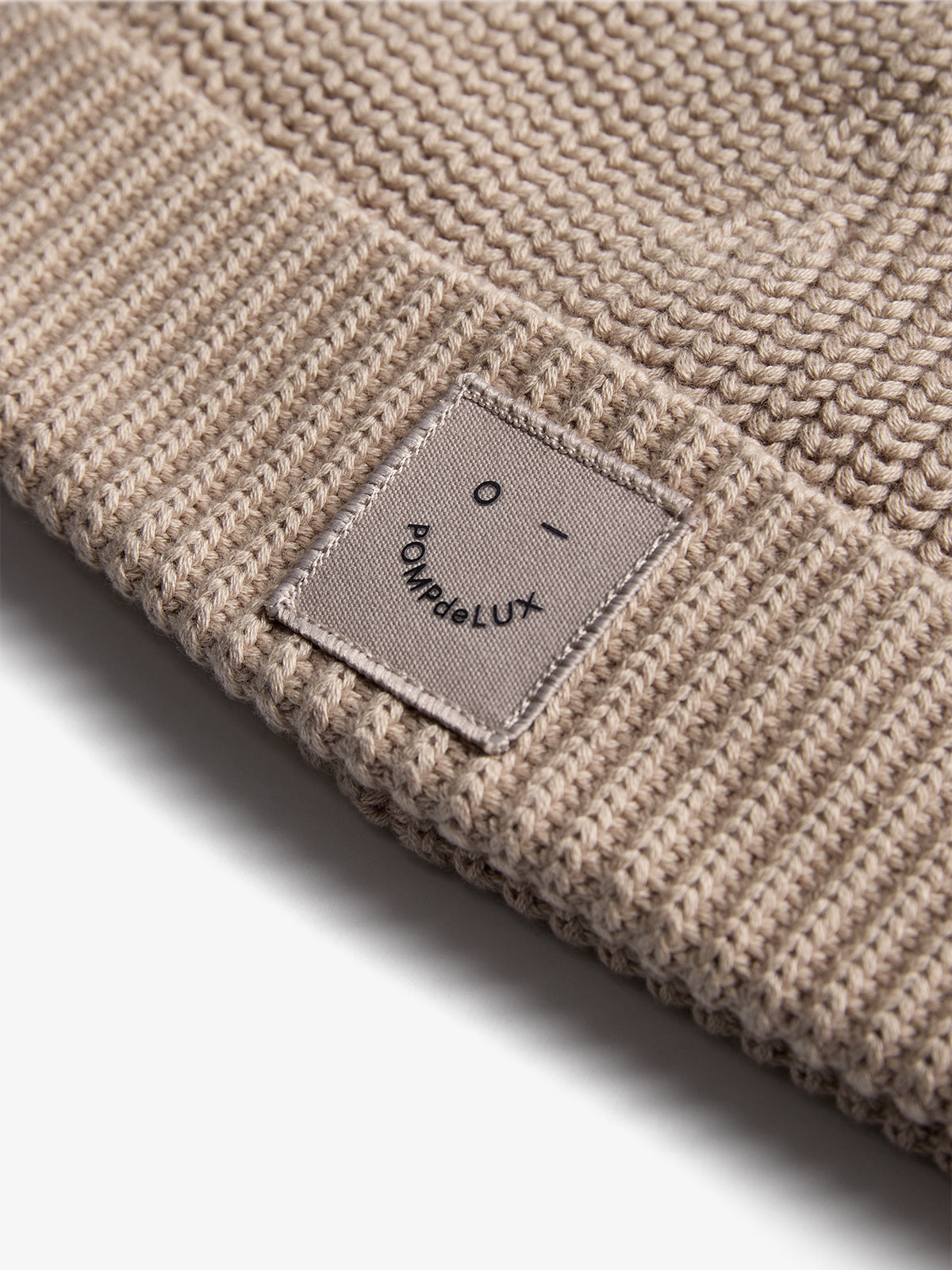 Strikket beige hue ligger fladt med et label på kanten. Label viser et enkelt ansigt og teksten "POMPdeLUX". Huen ligger på en lys baggrund.