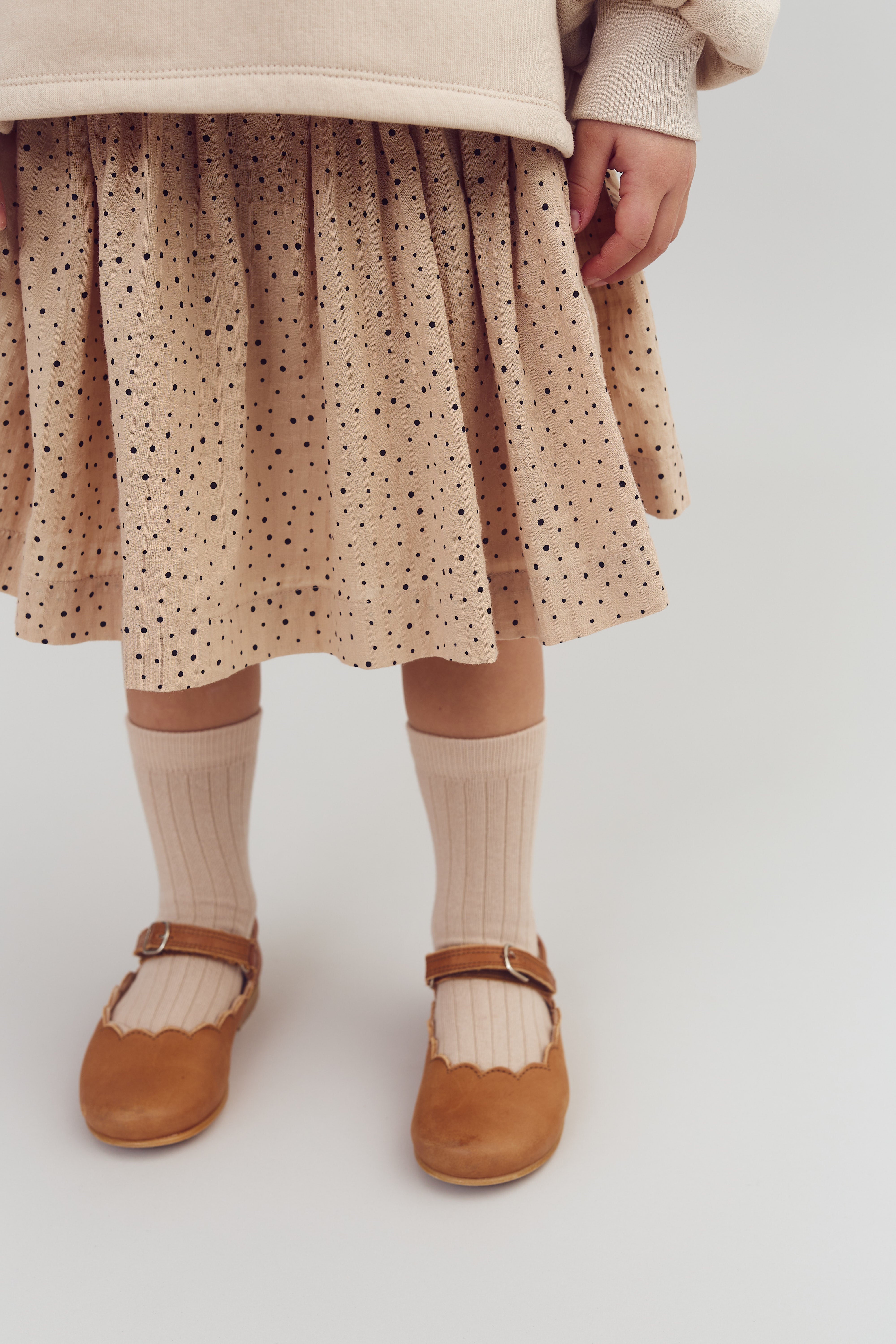 A child stands wearing a beige polka dot dress, cream socks, and brown Mary Jane shoes against a simple white background.