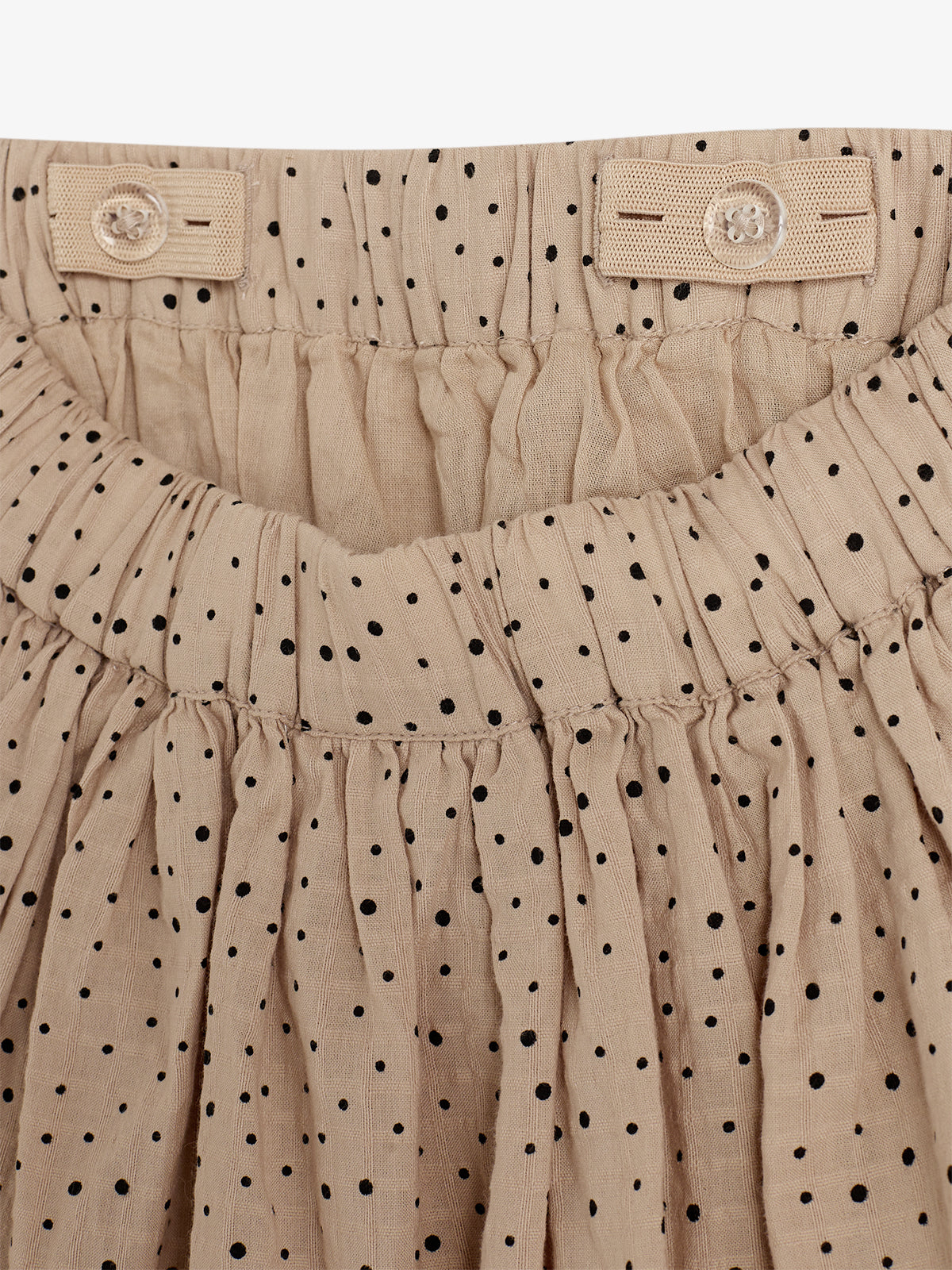 A beige gathered fabric skirt with black polka dots is shown with an elastic waistband and button details, emphasizing texture and design in a close-up view.