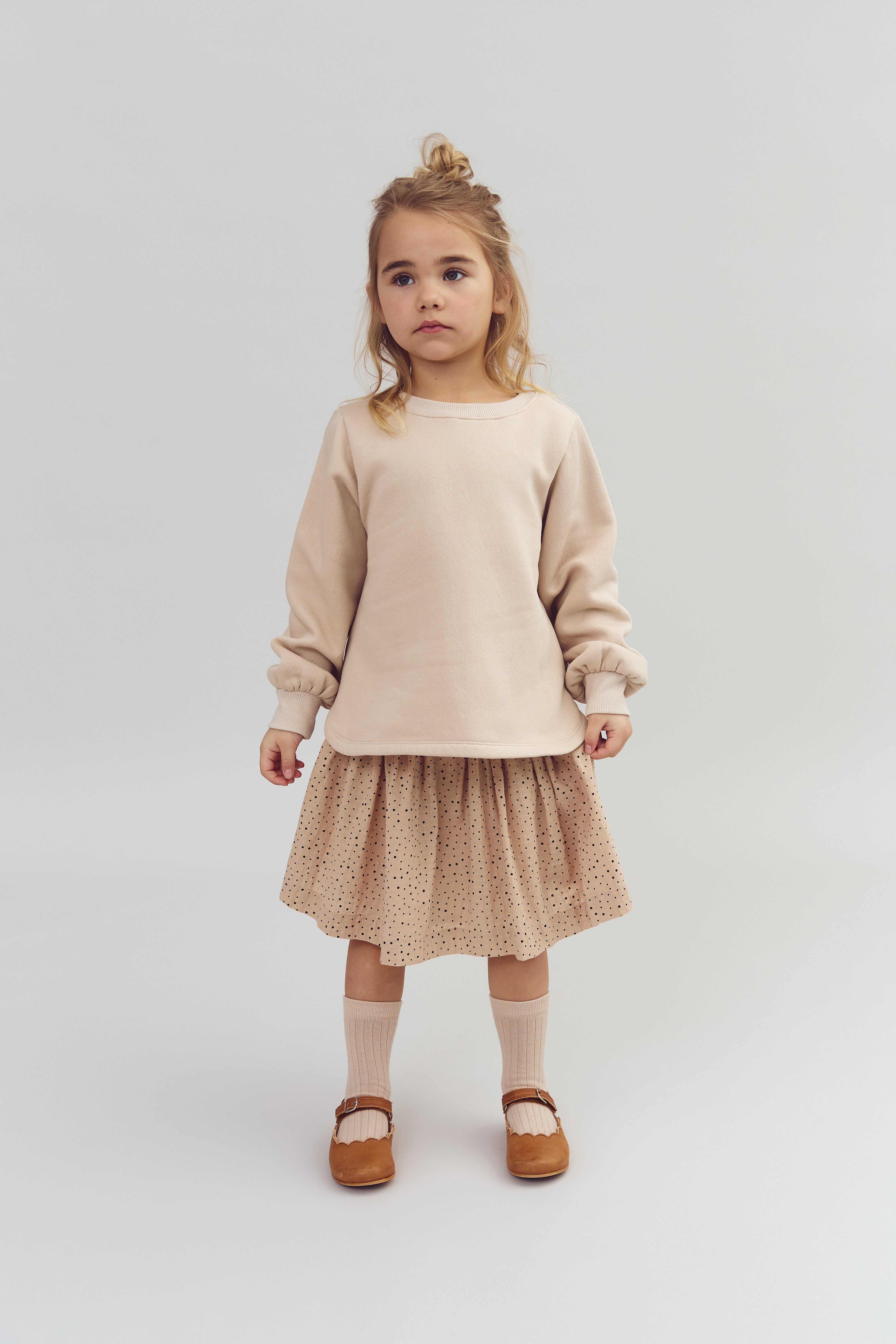 A child stands still wearing a beige sweater, a polka dot skirt, and brown shoes with socks, against a plain light gray background.