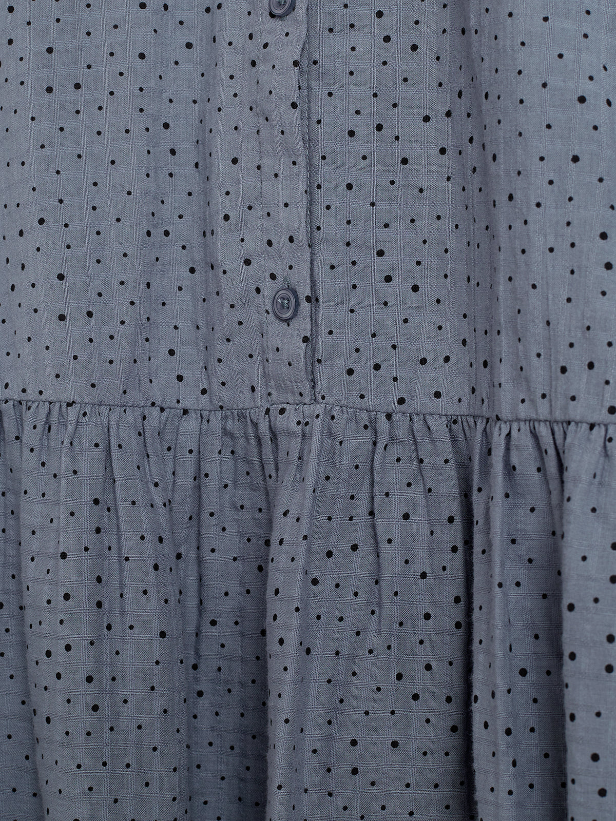 Gray fabric with small black polka dots forms a buttoned-up shirt. Ruffles are visible below the buttons, giving a textured appearance. The background is simply part of the fabric.