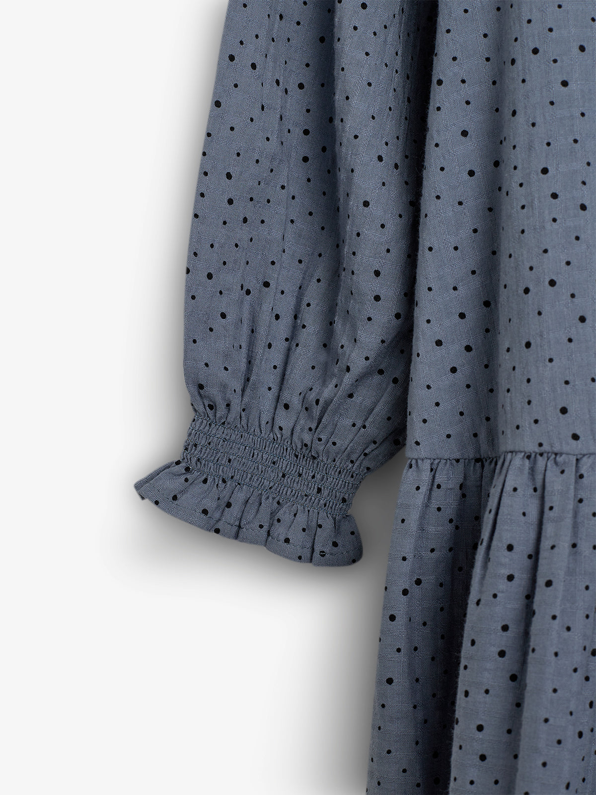 The image features a blue fabric with black polka dots, focusing on a sleeve with elasticized cuffs and a gathered, ruffled hem, set against a plain white background.