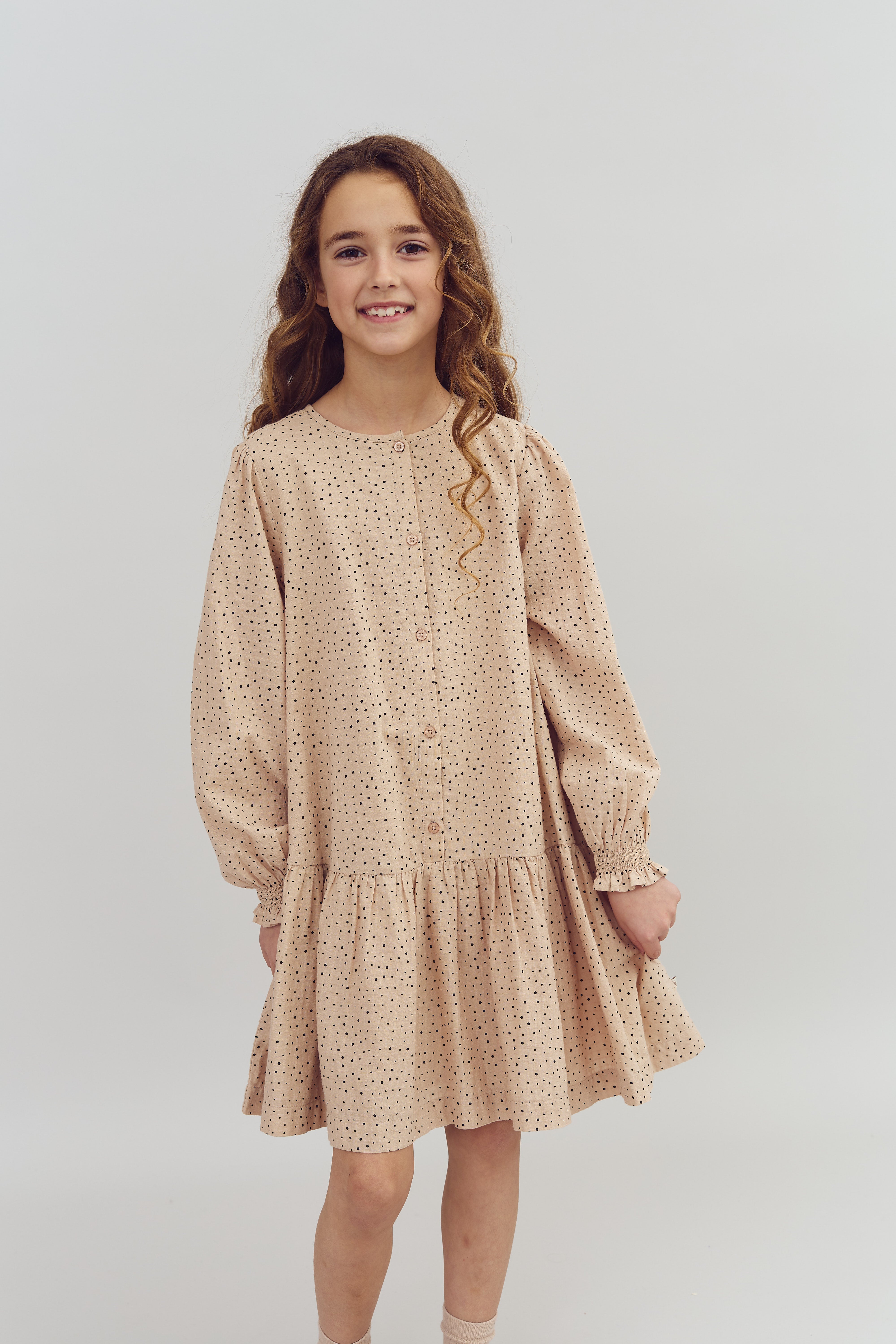 Pige smiler iført beige kjole med sorte prikker. Hun har krøllet hår. Baggrunden er en ensfarvet, lys væg.