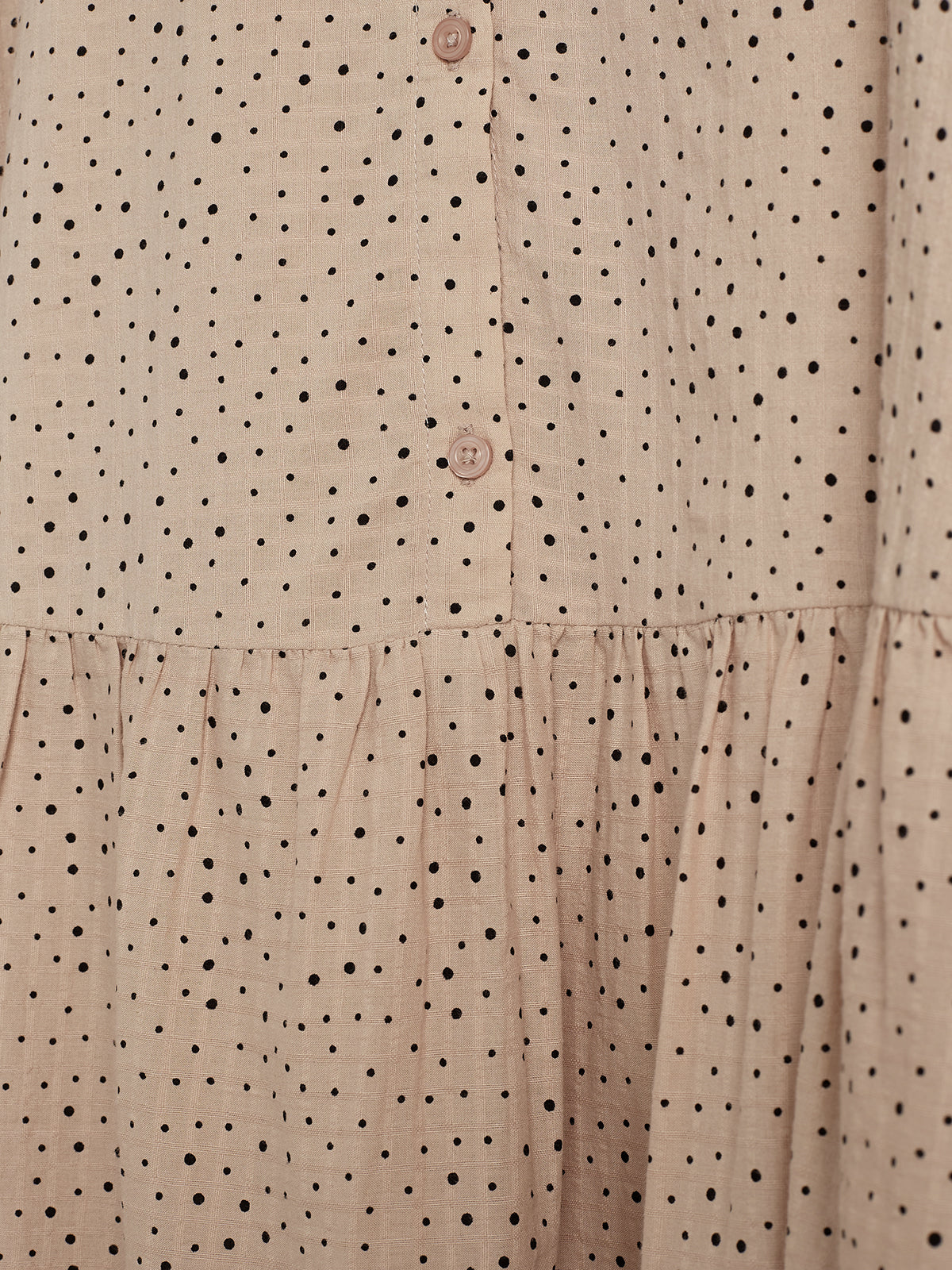 Beige stof med små sorte prikker, knappet med to brune knapper. Stoffet er let rynket ved kanten, hvilket skaber et let bølgende udtryk.