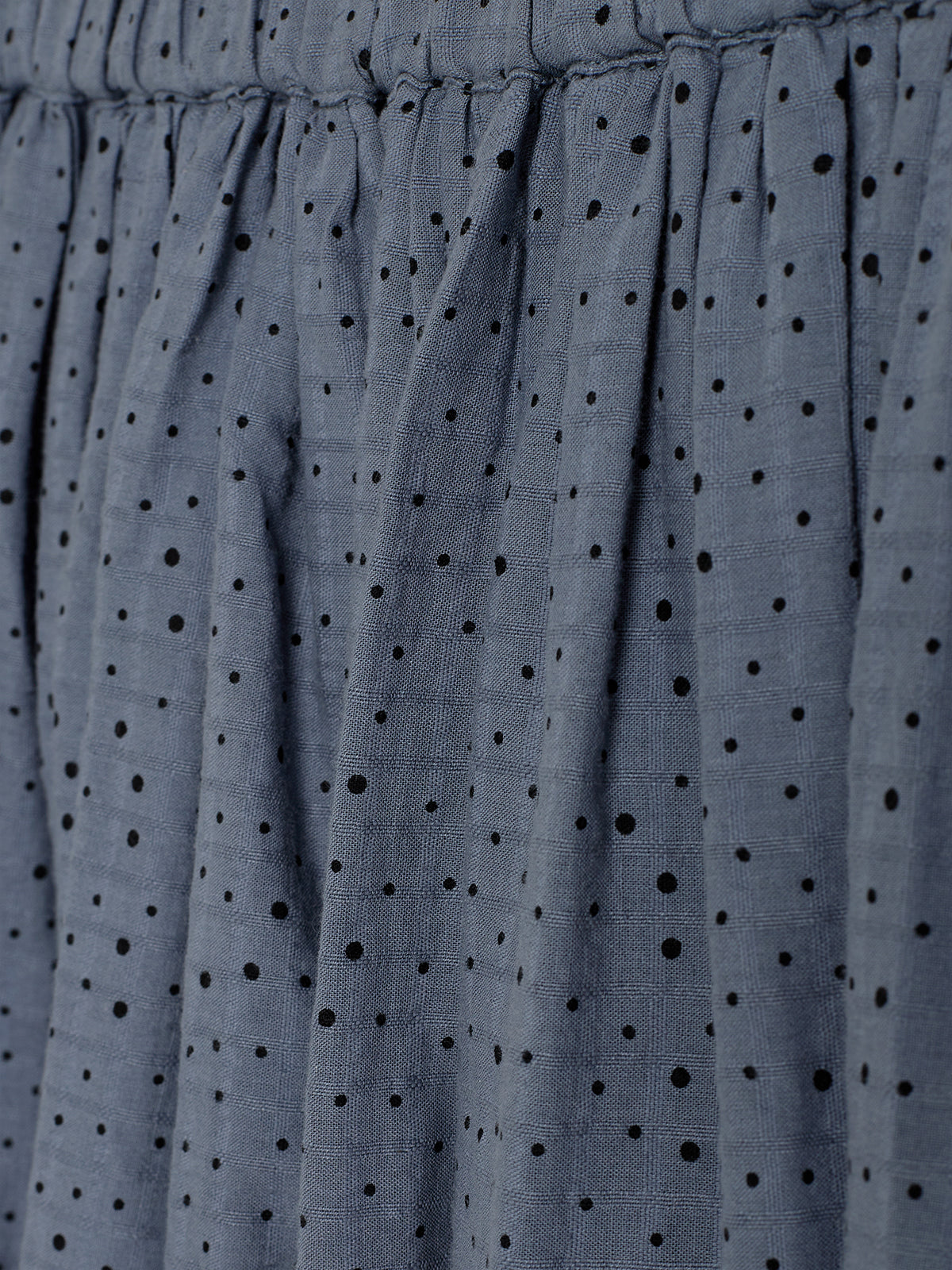 The fabric features tightly gathered pleats with a pattern of small black polka dots, set against a textured, gray-blue background.