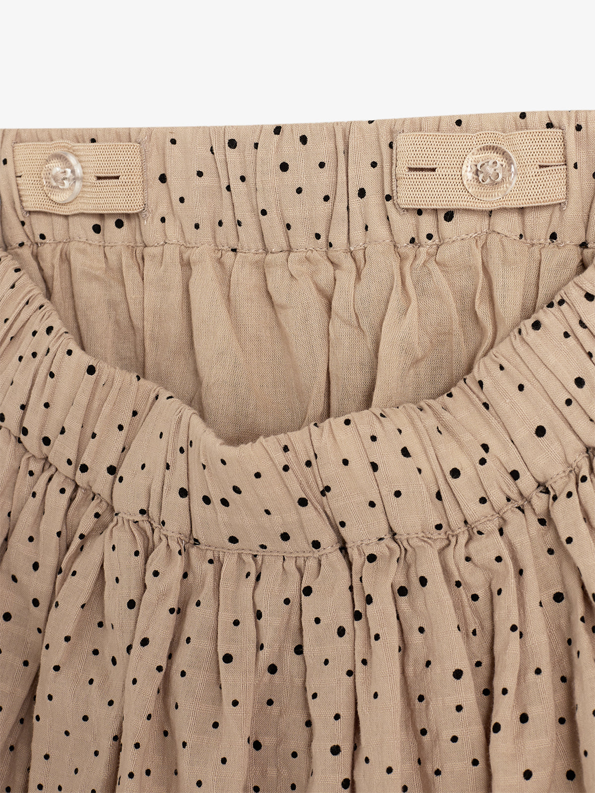 Beige fabric skirt, featuring a gathered waistband with adjustable button tabs, adorned with small black polka dots throughout, creating a textured, patterned design in a neutral setting.