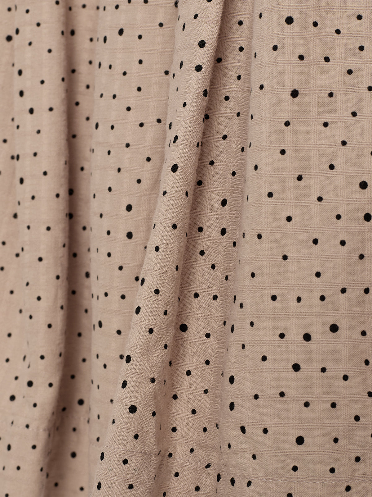 Beige fabric drapes feature small black polka dots in an irregular pattern, creating a textured appearance. The fabric hangs against a plain background, emphasizing its soft folds and grid-like weave.