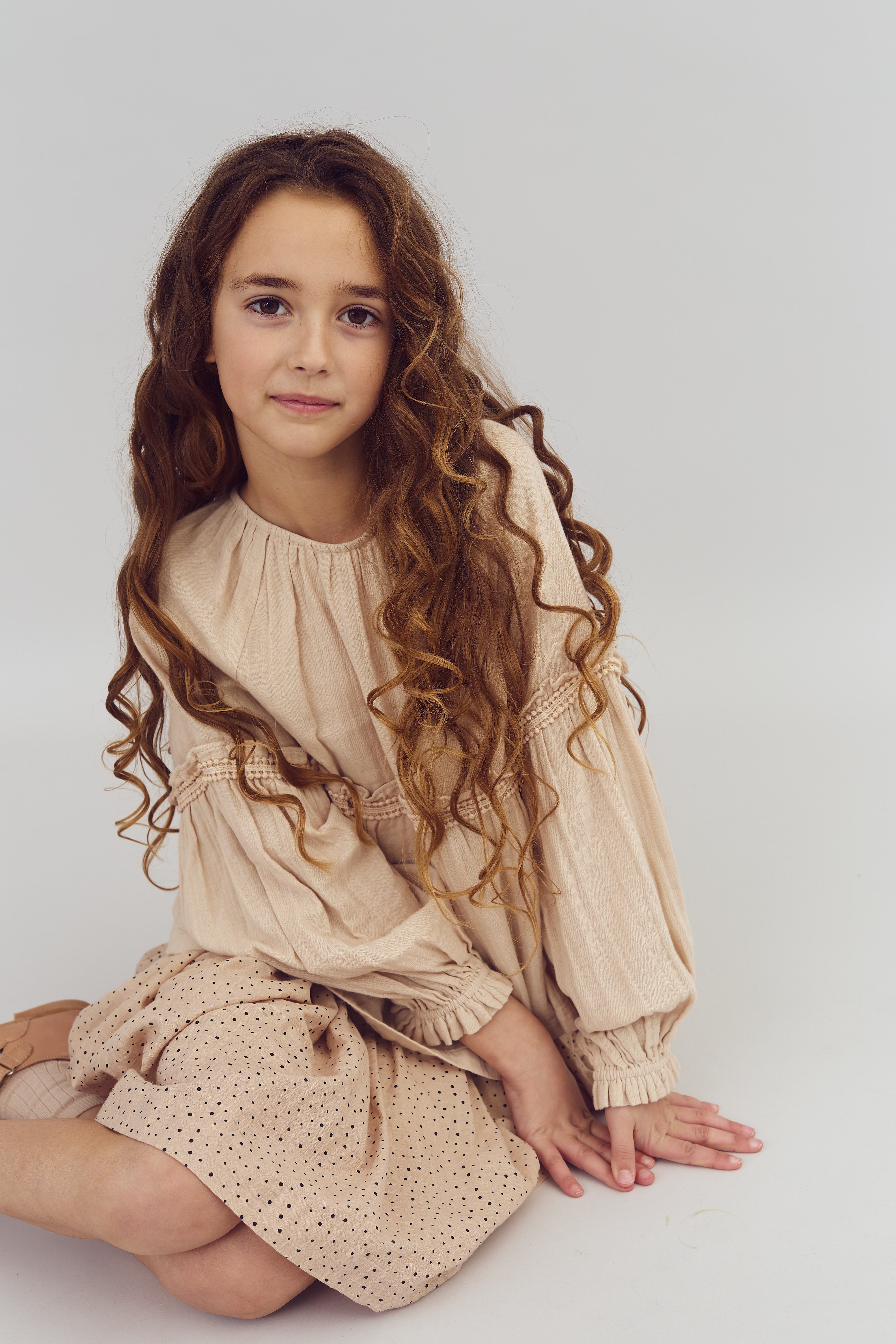 A young girl with long, curly hair sits on the floor wearing a beige dress with decorative sleeves in a neutral, indoor setting.