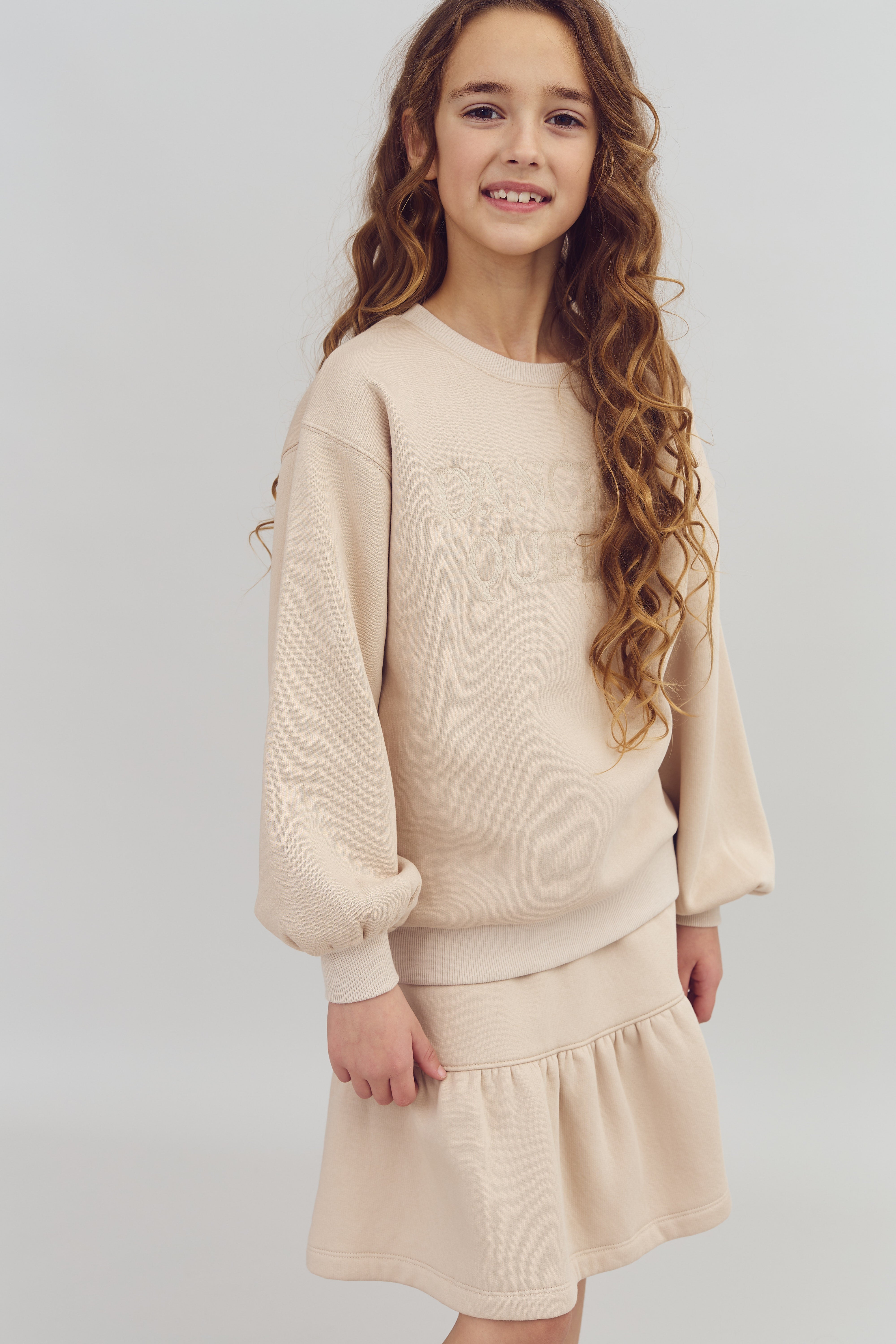 Pige smiler iført en lys beige sweater med teksten "DANCE QUEEN," matcher en nederdel, står mod en neutral baggrund.