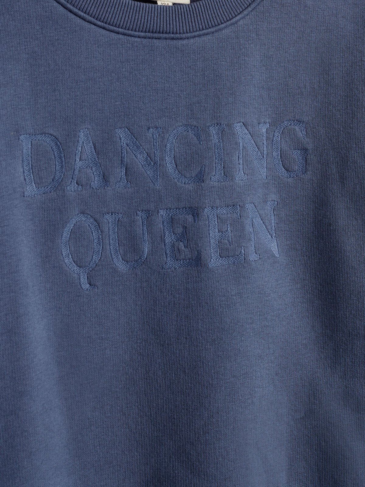 Tekst på en blå sweatshirt viser ordene "DANCING QUEEN" i hævet, matchende blå skrift. Trøjen hænger i et neutralt lys uden synlige baggrundsdetaljer.