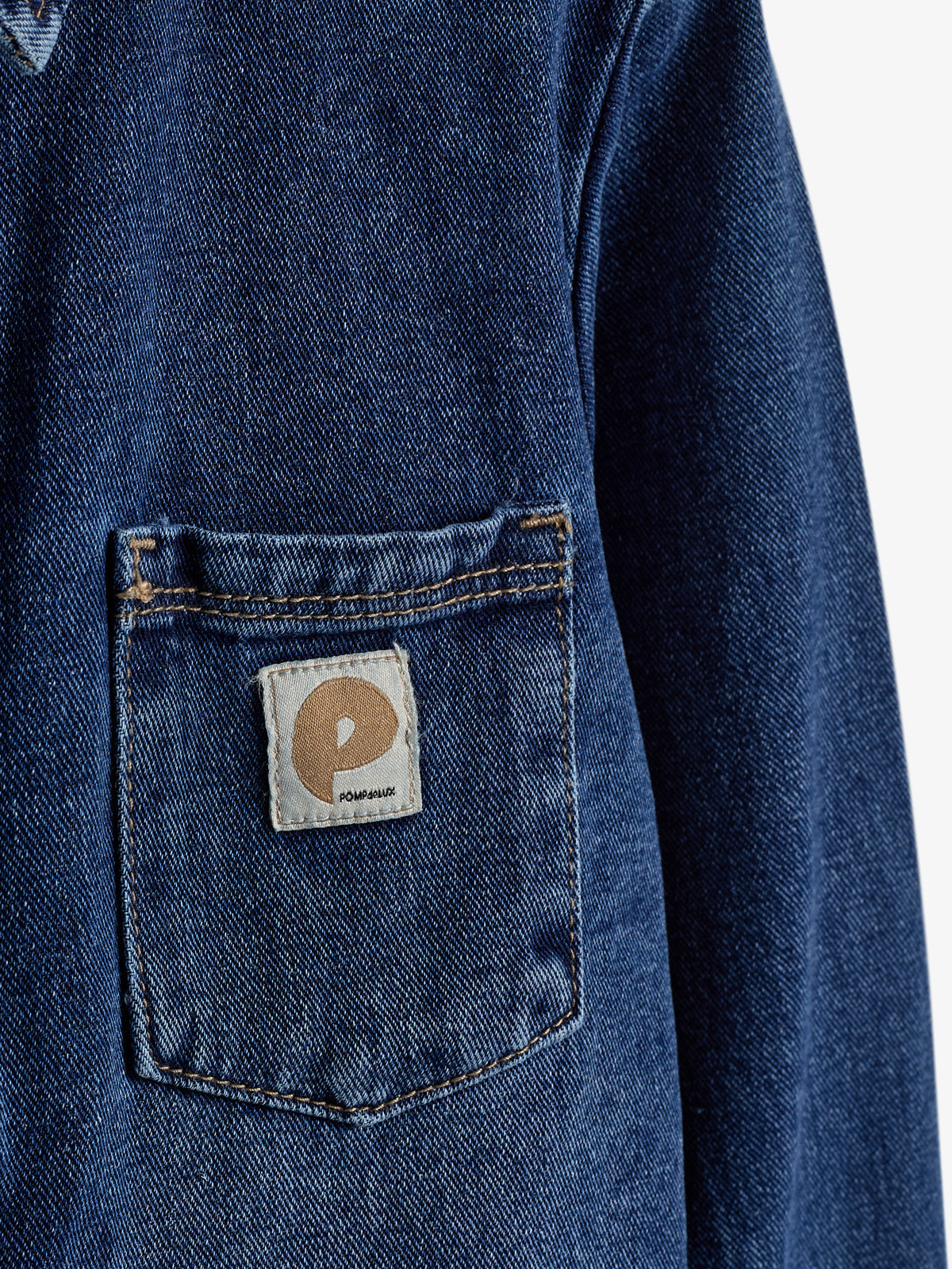 Denimjakke; har en baglomme med et firkantet mærketikette med bogstavet "P" og teksten "POMPAUX"; tæt, neutral baggrund.