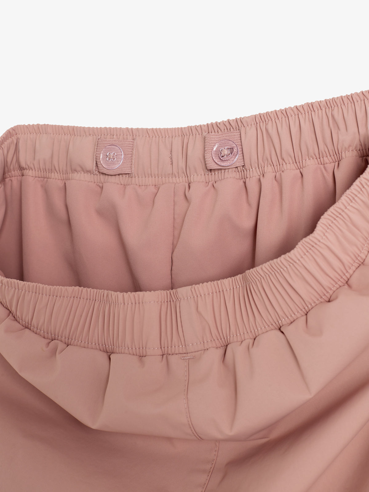 Bukser med rosa elastisk talje og justerbare knapdetaljer ses oppefra, fokus på taljebåndet. Stoffet er glat og let rynket, omgivende hvid baggrund.